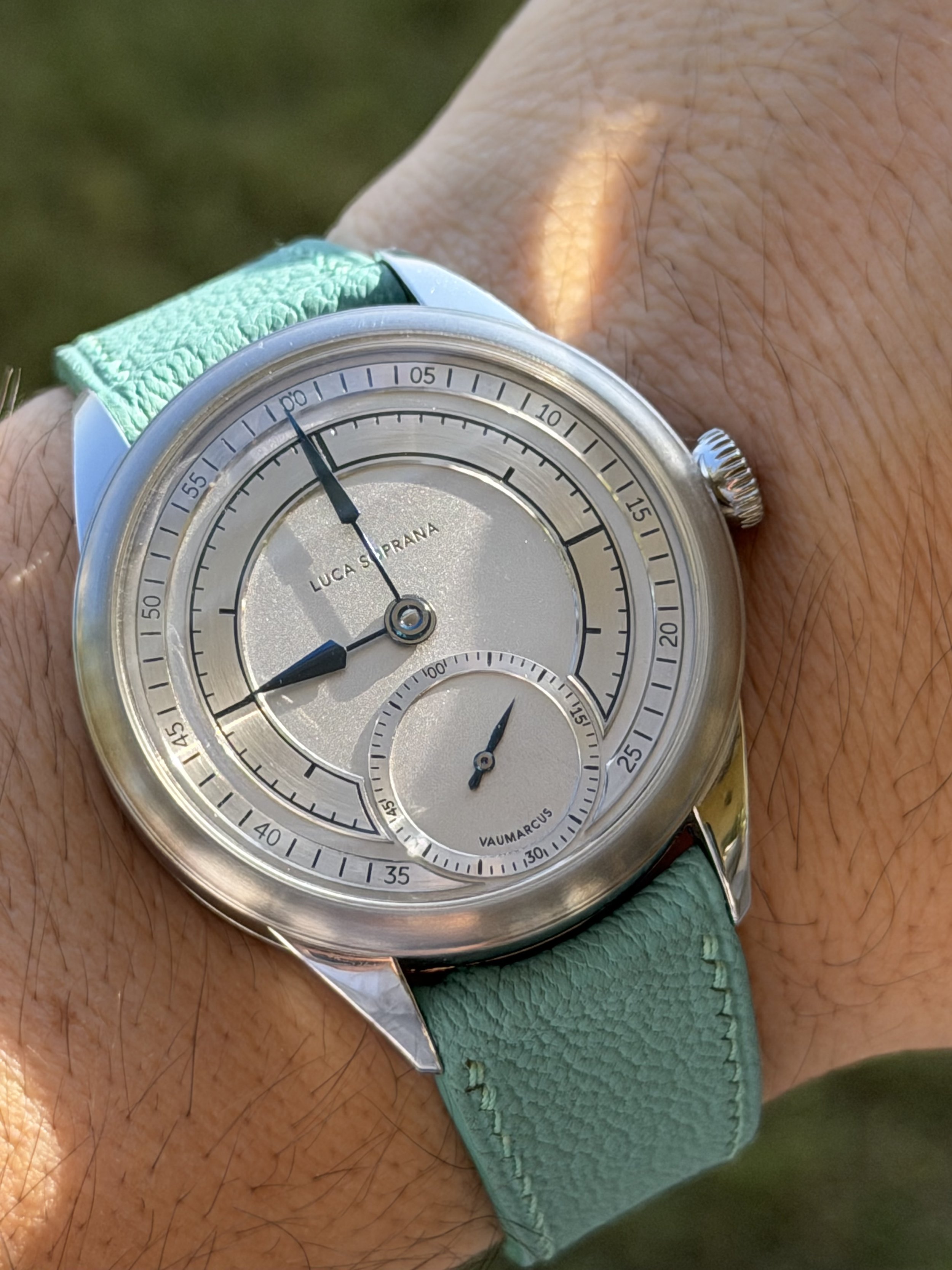 Close-up of a wristwatch with a silver face, dark hands, and a small subdial, worn on a wrist with a light green strap. luca soprana