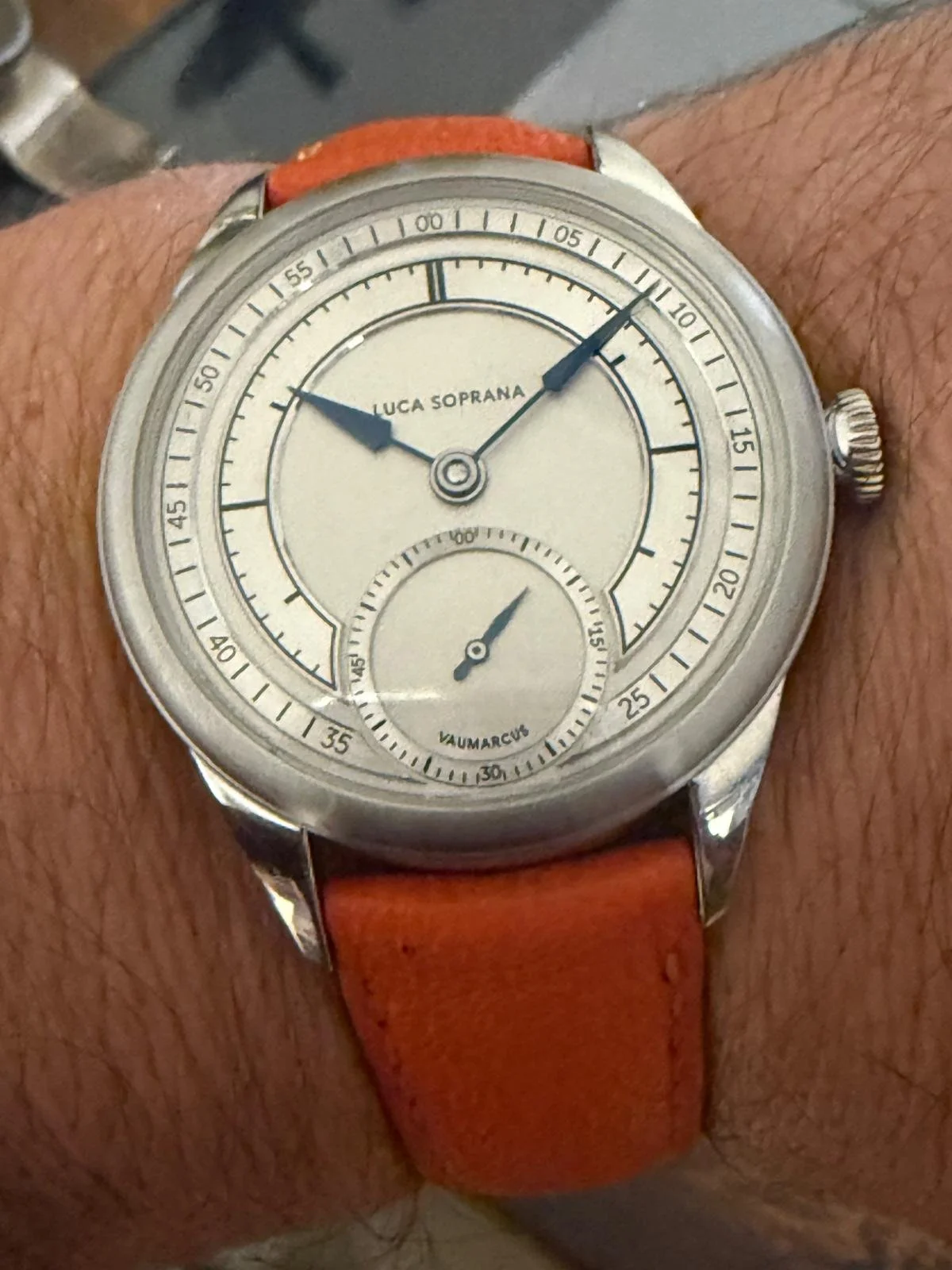 A wristwatch with a cream dial, black hour and minute hands, and a small seconds subdial at 6 o'clock. The watch face has the names LUCA SOPRANA and VAUMARCH. It features a red leather strap.