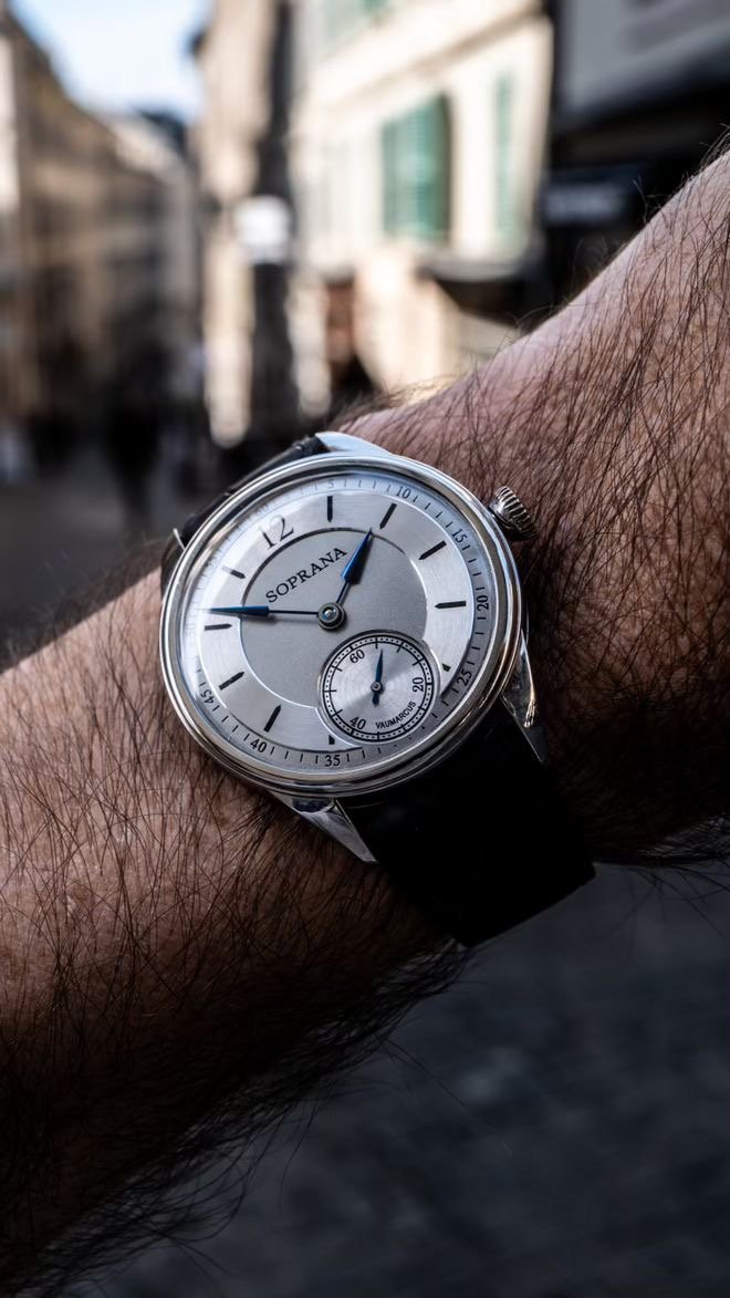 Close-up of a wristwatch on a man's wrist showing the time as 2:44, with buildings and a street in the blurred background.