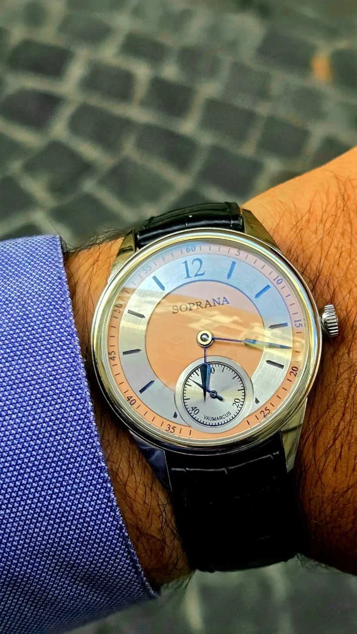 A wristwatch with a round face showing the time, worn on a person's left wrist, with a black leather strap and a silver case, against a blurred cobblestone background.