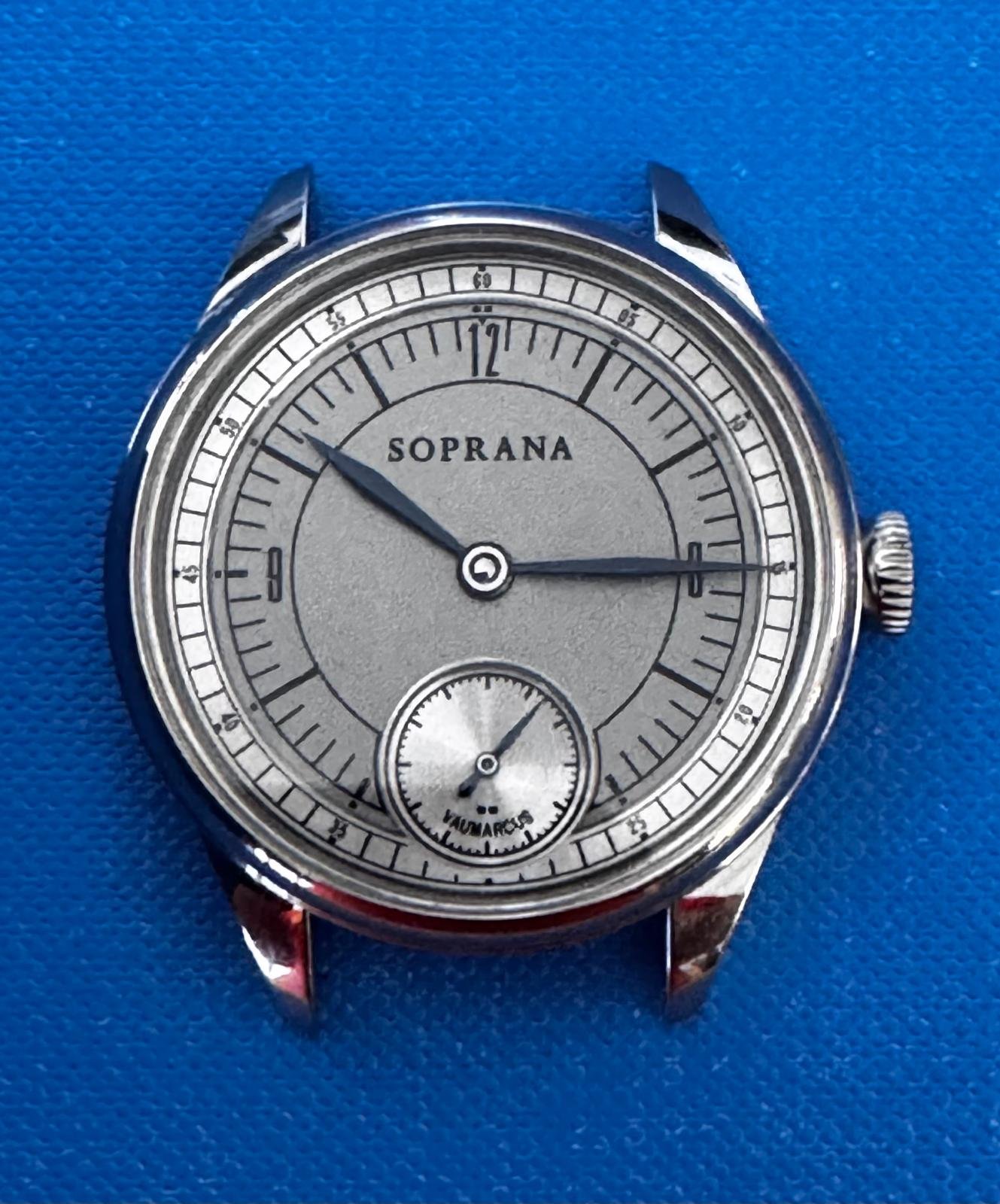 A vintage wristwatch with a silver dial, black hour and minute hands, and a small seconds sub-dial at the 6 o'clock position. The brand name "SOPRANA" is printed on the dial. The watch has a stainless steel case and is placed on a blue textured surfa