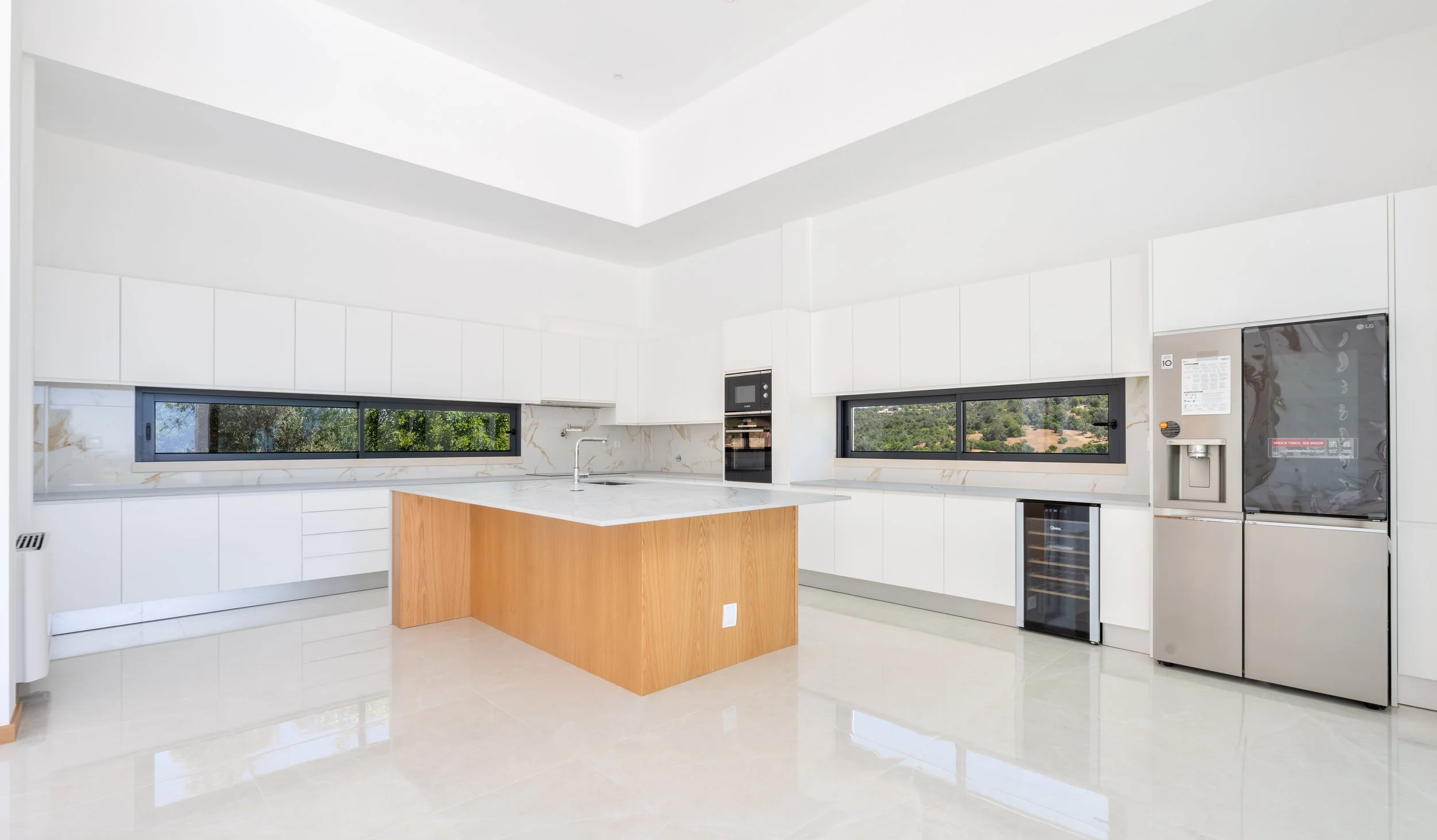 Cozinha moderna com armários brancos, ilha de cozinha de madeira, janelas amplas com vista para a natureza, geladeira prateada e eletrodomésticos embutidos.