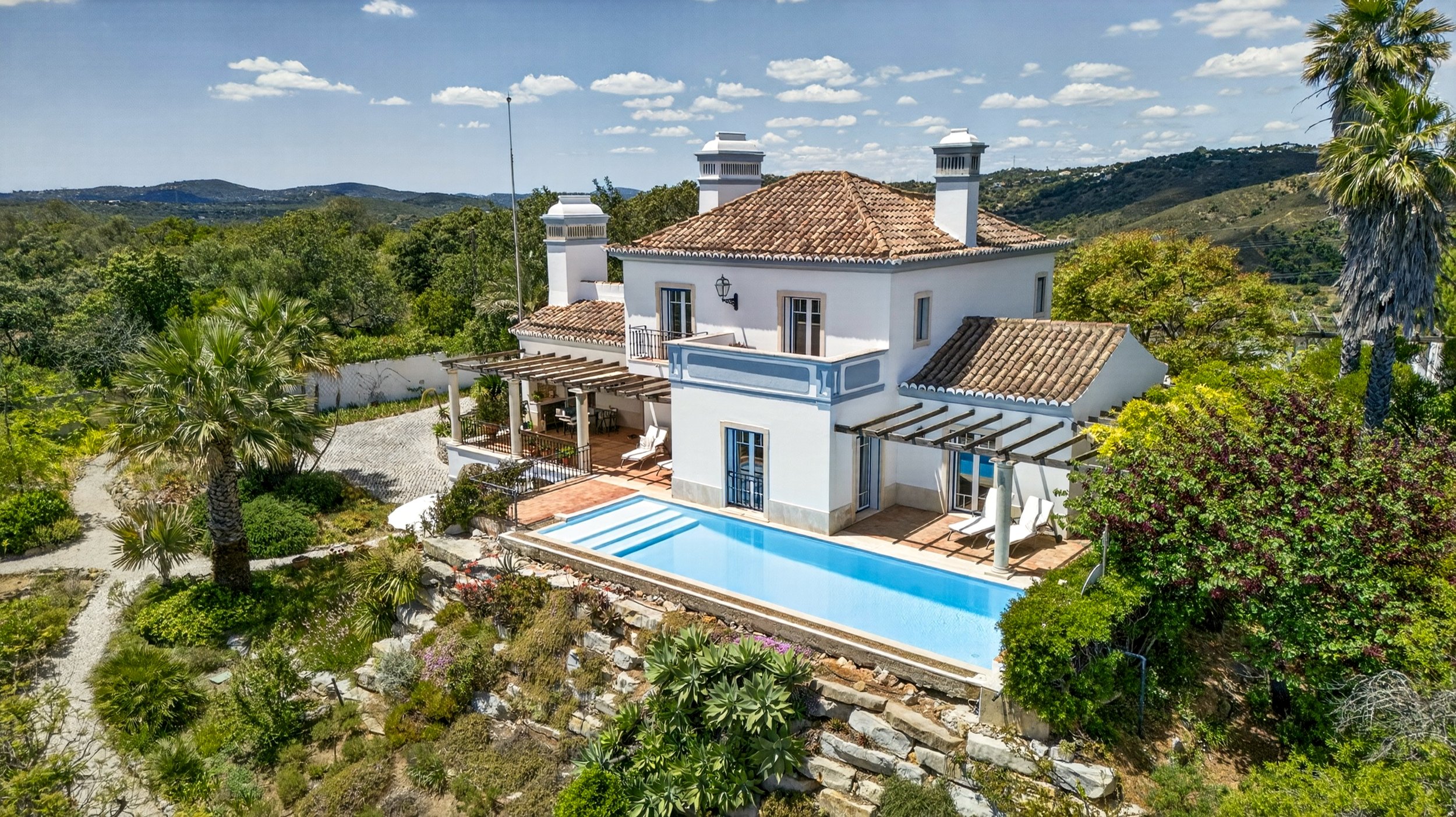 Casa luminosa com telhado de telhas cerâmicas, varanda com pérgula de madeira, piscina e jardim com árvores e plantas verdes, vista para áreas rurais e montanhas ao fundo.
