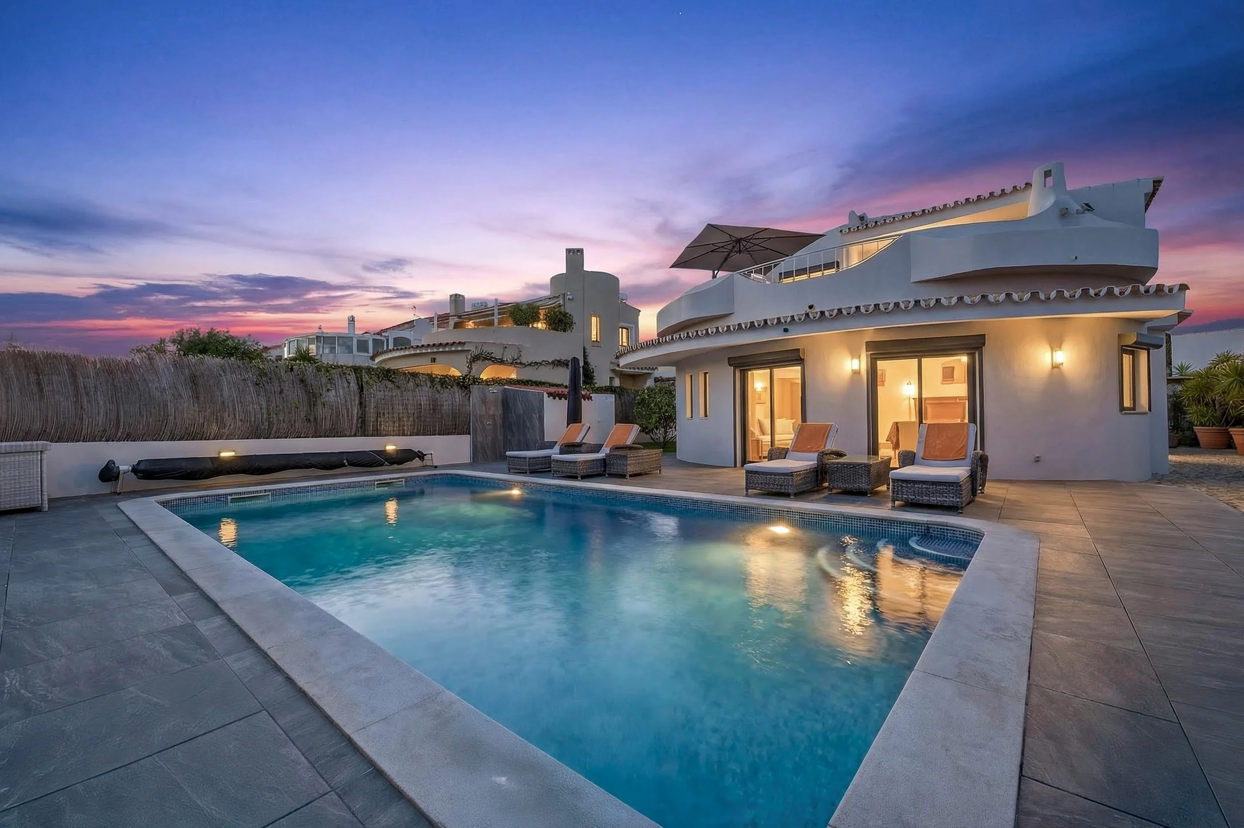 Casa branca com telhado de clay tiles, varanda com guarda de vidro, janela iluminada, piscina ao ar livre com cadeiras e sombreiro em um ambiente ao pôr do sol.