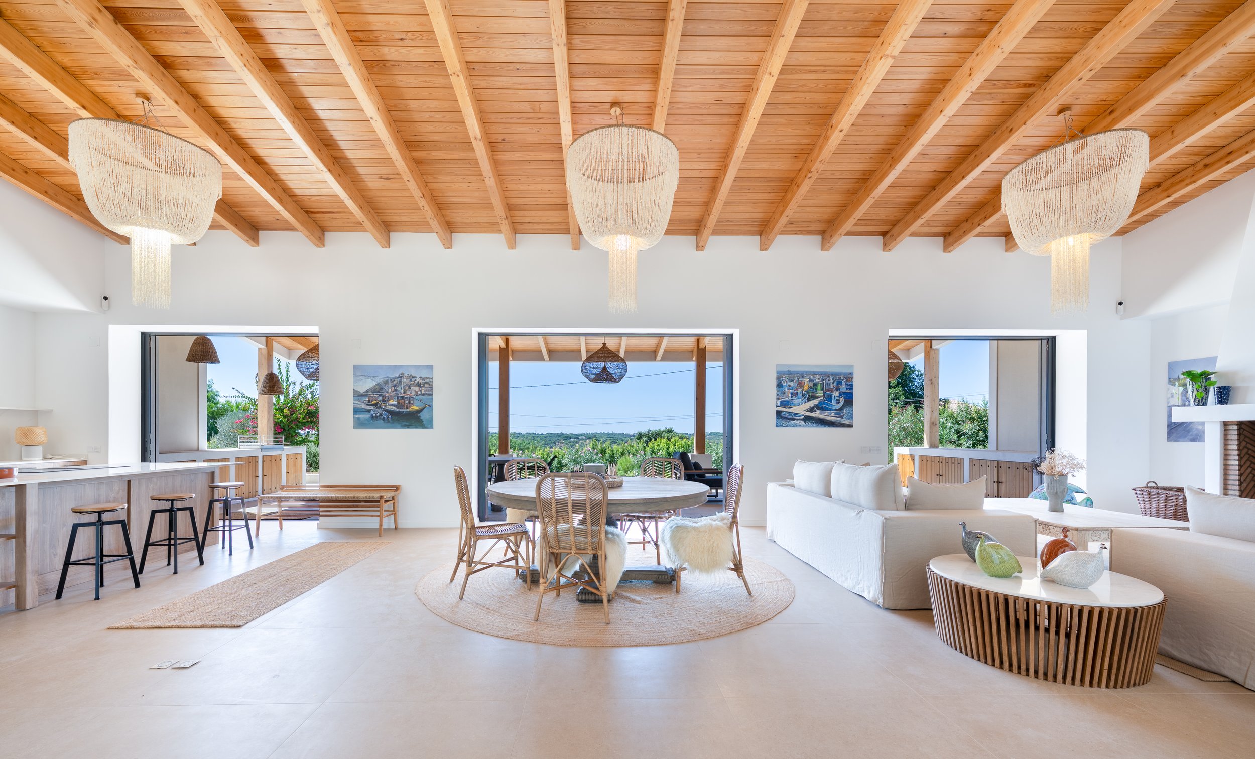 Living room interior with a wooden ceiling, chandelier lighting, large windows overlooking the swimming pool and outdoor area, light fabric furniture and natural-fibre decorative elements.