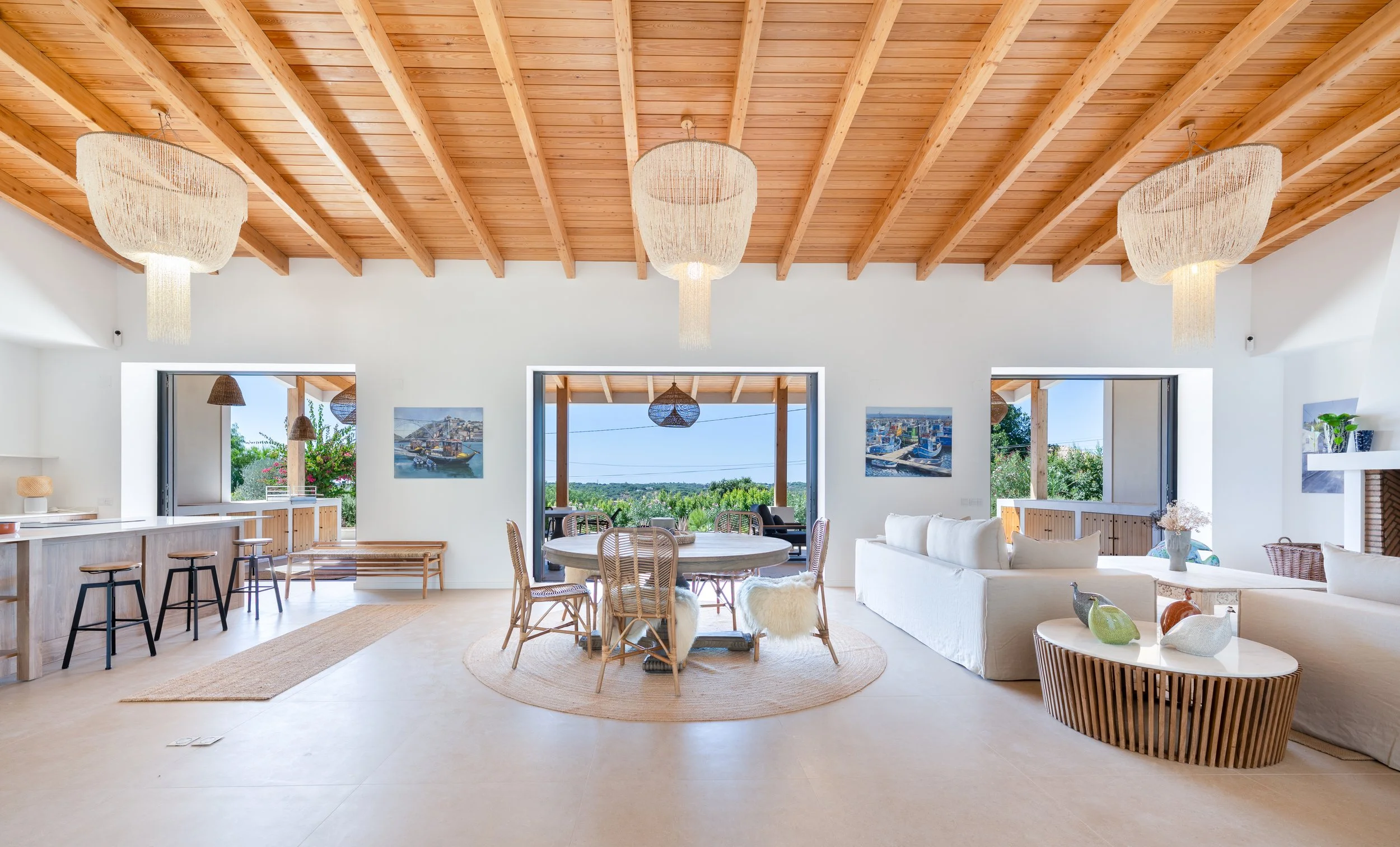 Interior de sala de estar com teto de madeira, luminárias de chandelier, janelas grandes com vista para piscina e área externa, móveis de tecido claro e decorativos de fibra natural.