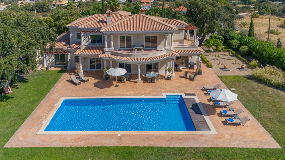 Uma casa grande com varanda, área de piscina ao ar livre, cadeiras de sol, guarda-sóis, mesas e cadeiras na área da piscina, jardim bem cuidado ao redor, árvores e vegetação ao fundo.