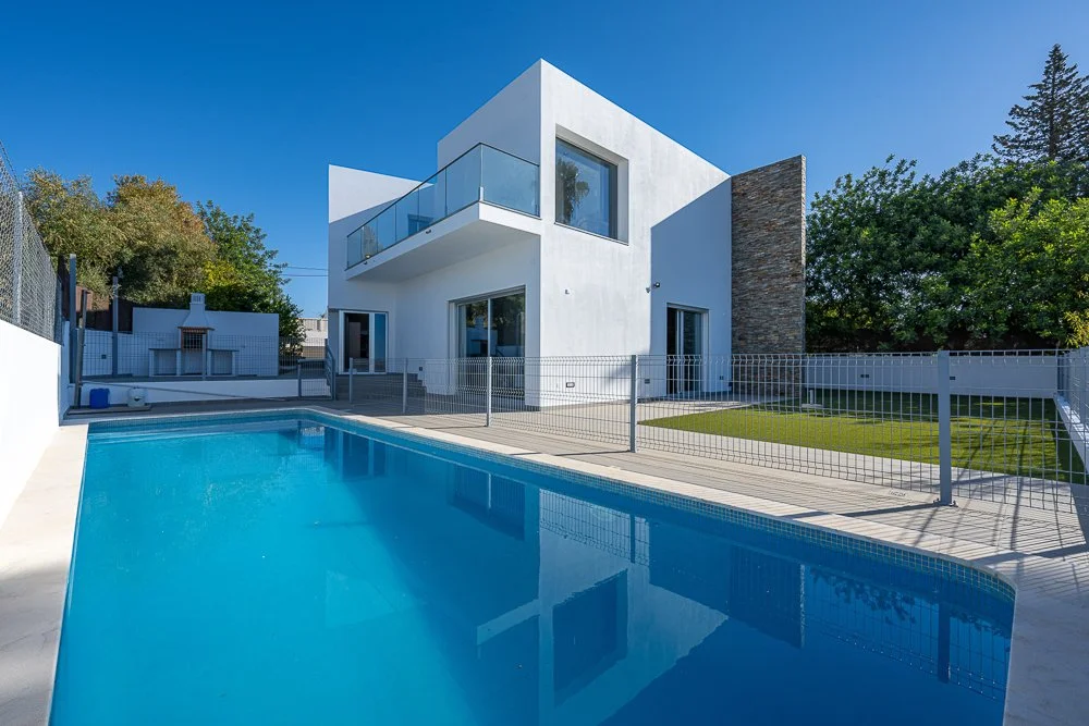 Casa moderna branca com piscina e jardim, cercada por uma cerca de metal, sob céu azul e árvores ao fundo.