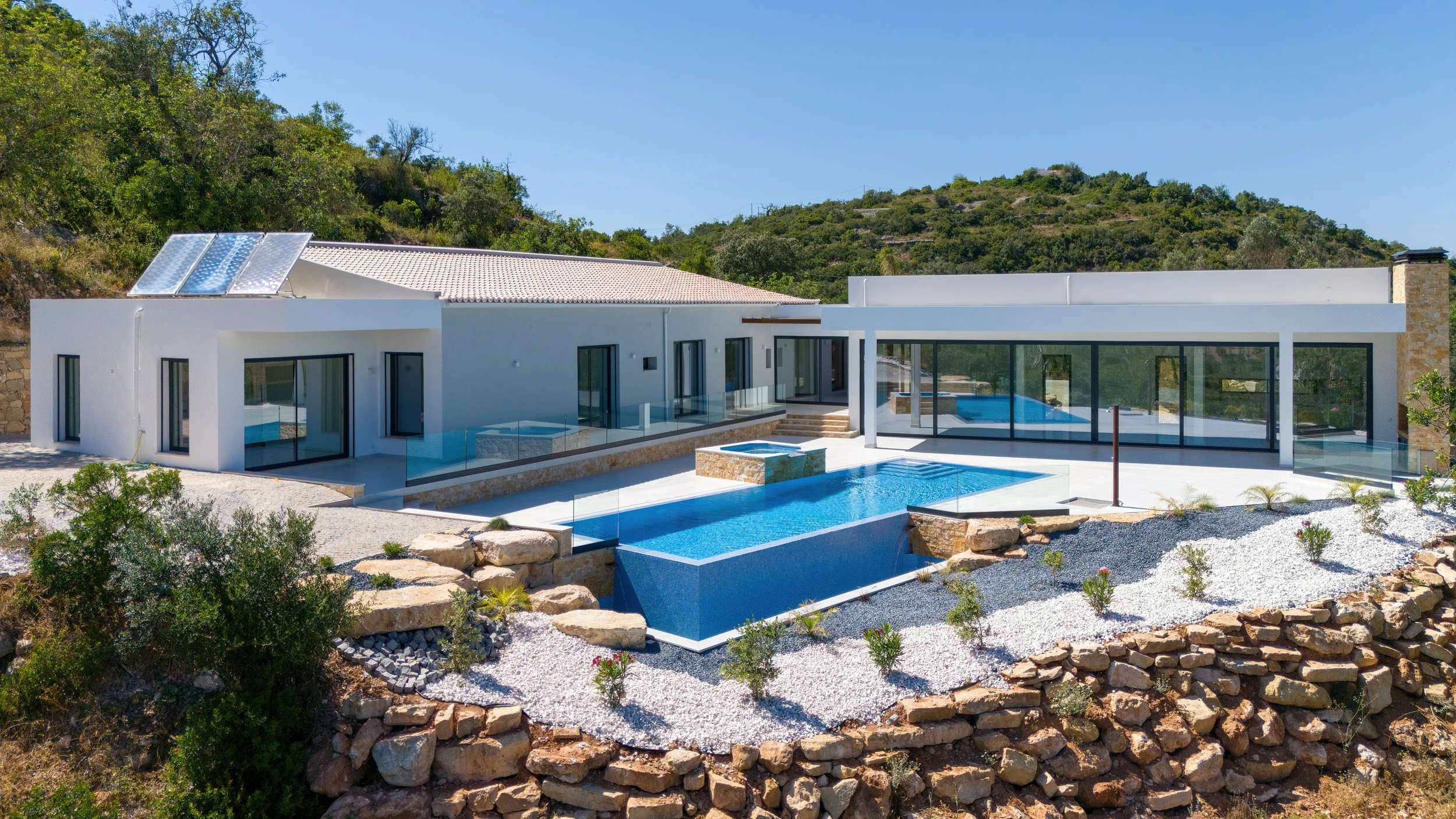 Casa moderna com piscina e jardim em terreno de relevo, cercada por vegetação e colinas ao fundo.
