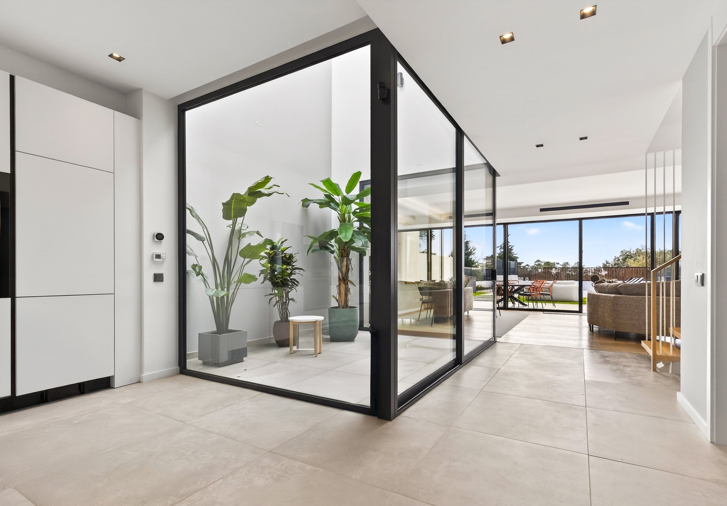 Ambiente moderno com paredes brancas, piso de cerâmica, plantas internas, sala de estar com móveis confortáveis, grande janela com vista para o exterior ensolarado.