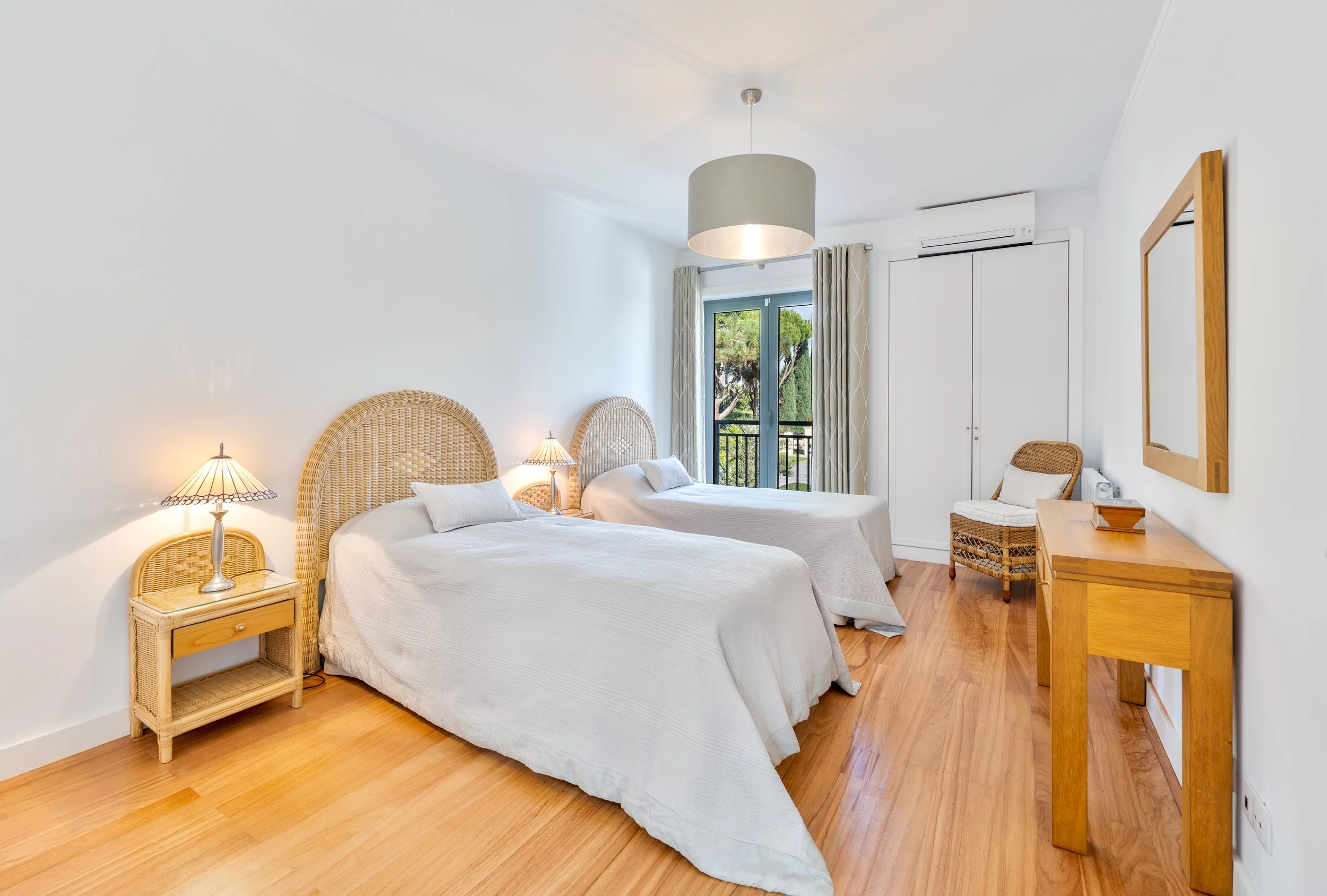 Quarto com duas camas de casal com cabeceiras de fibra natural, uma mesinha de cabeceira com abajur, uma cadeira de fibra natural, um espelho de madeira na parede e uma janela com cortinas, com vista para árvores.