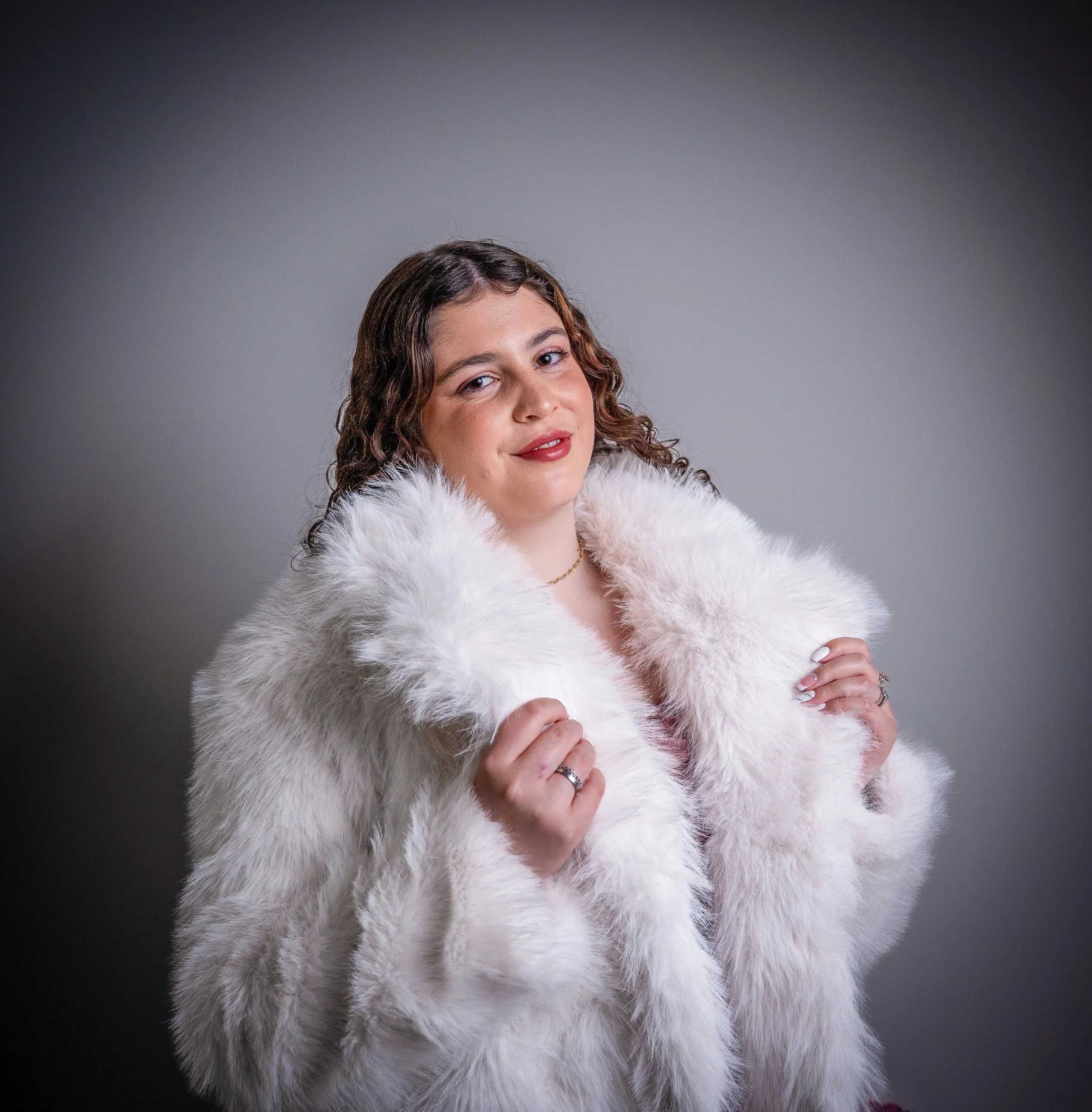 Retrato de uma mulher com cabelo cacheado, usando blush, batom vermelho e vestido branco, com fundo cinza.