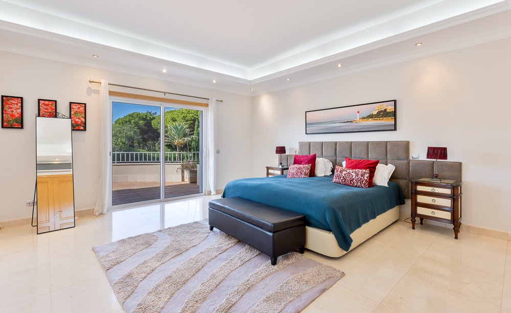 Quarto de casal com cama grande, cabeceira estofada, roupas de cama azul, almofadas vermelhas e brancas, duas mesinhas de cabeceira com luminárias vermelhas, tapete lateral, porta de vidro que dá para uma varanda com vista para árvores, quadro na par