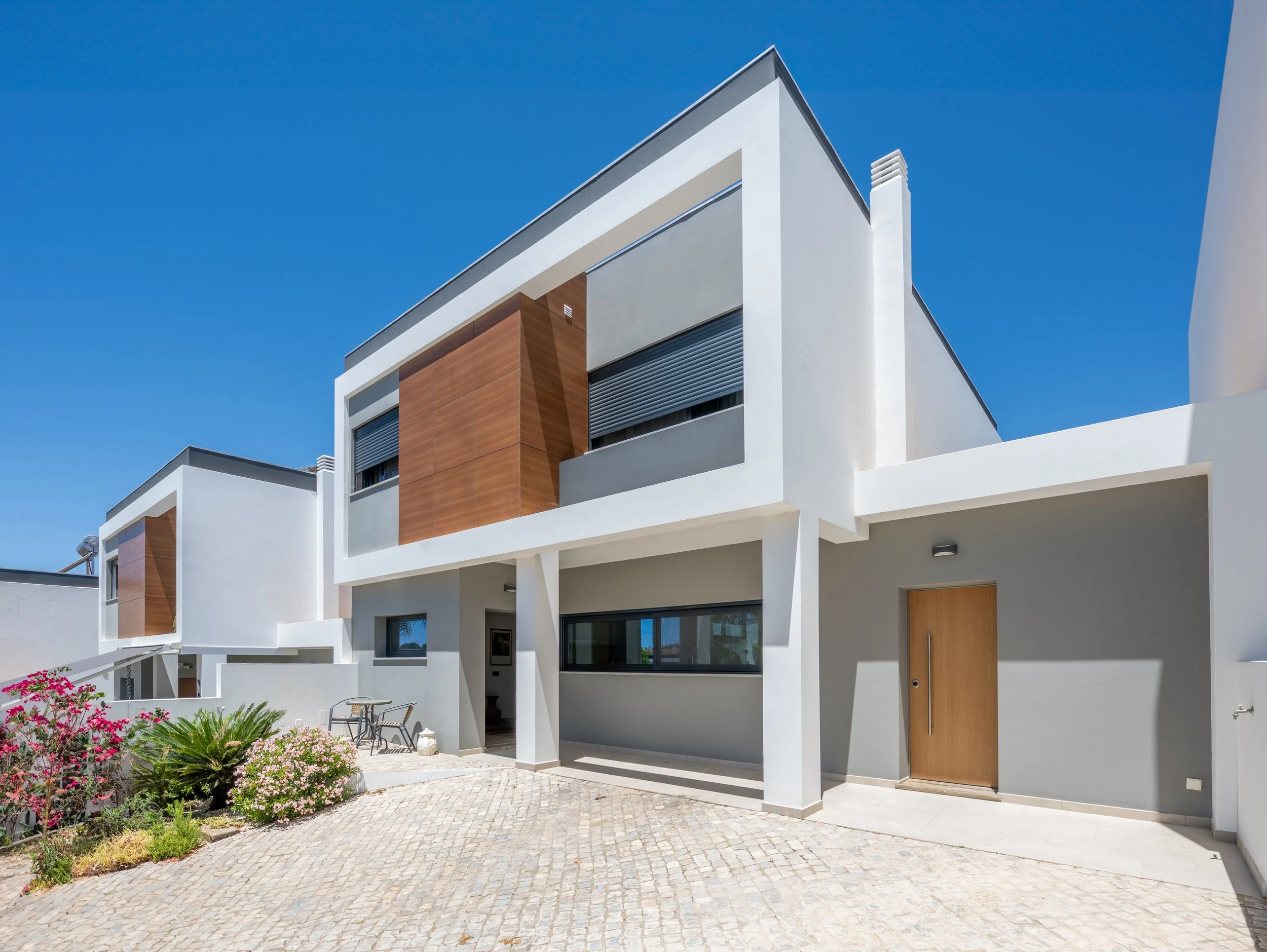 Prédio moderno com arquitetura contemporânea, fachada branca com detalhes em madeira, janelas grandes e porta de madeira, cercado por jardim com plantas e caminhos de pedra, sob céu claro e azul.