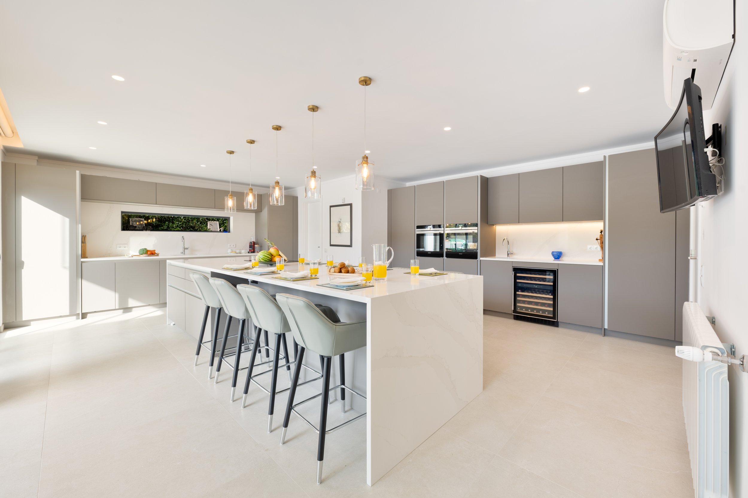 Cozinha moderna com ilha central, teto branco, iluminação pendente, armários cinza, televisão na parede, janelas altas, eletrodomésticos embutidos, bancada com frutas e sucos.