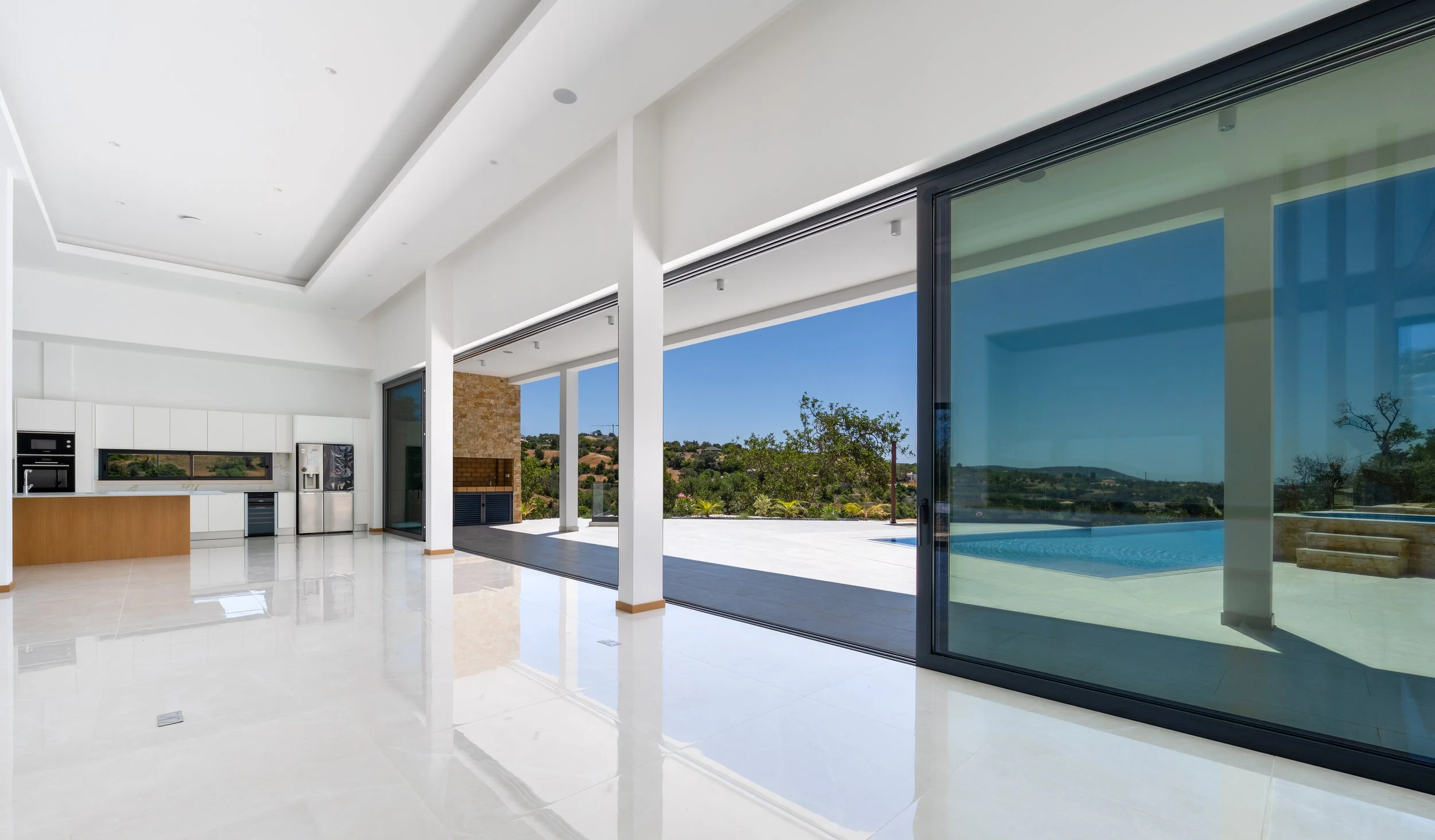 Interior de uma casa moderna com piso de mármore branco, cozinha branca com ilha de madeira, portas de correr de vidro que levam a uma piscina ao ar livre, vista para uma área com árvores e morros ao fundo.