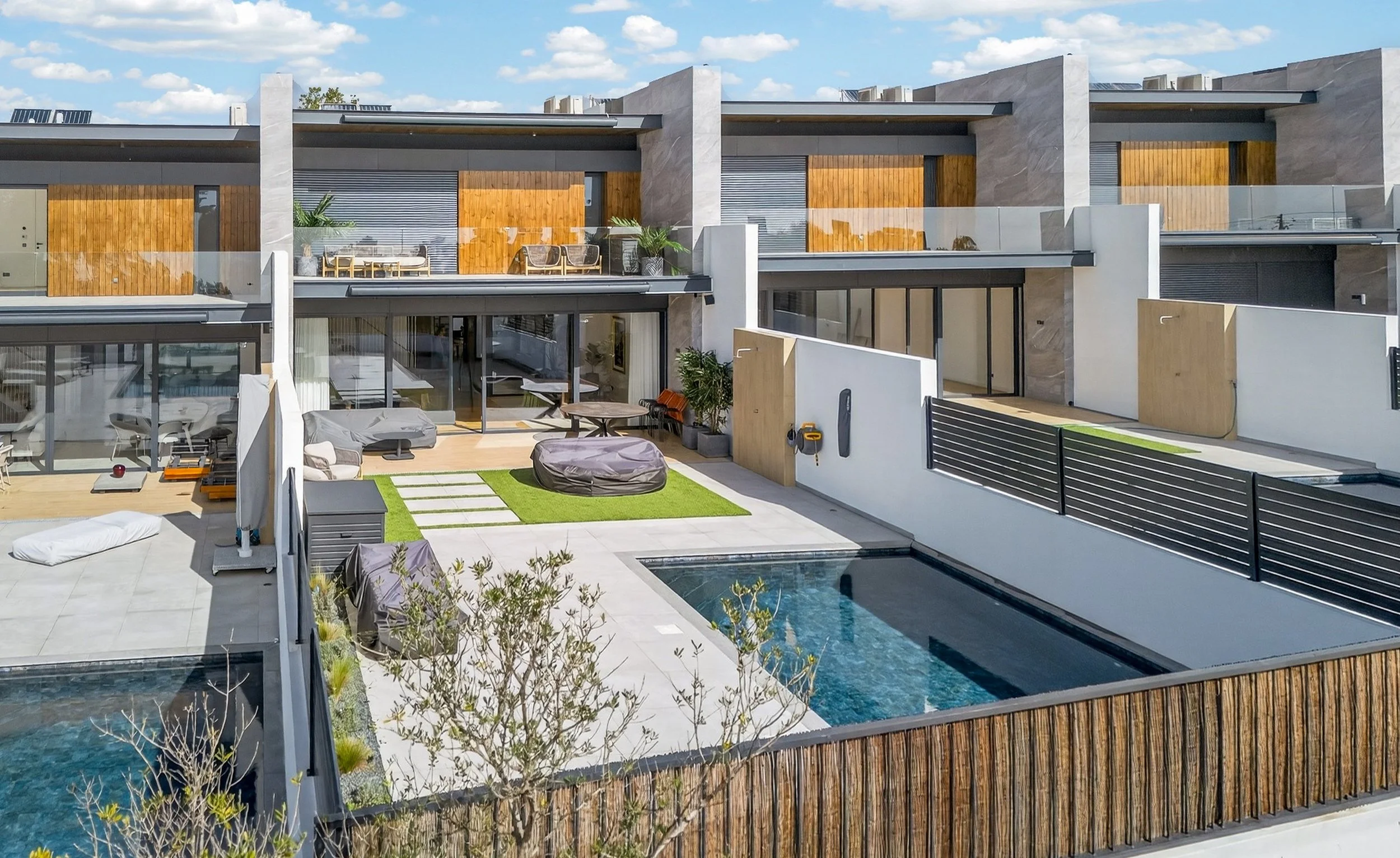 Complexo de apartamentos modernos com piscina, áreas externas com móveis e terraços com elementos de madeira e vidro.