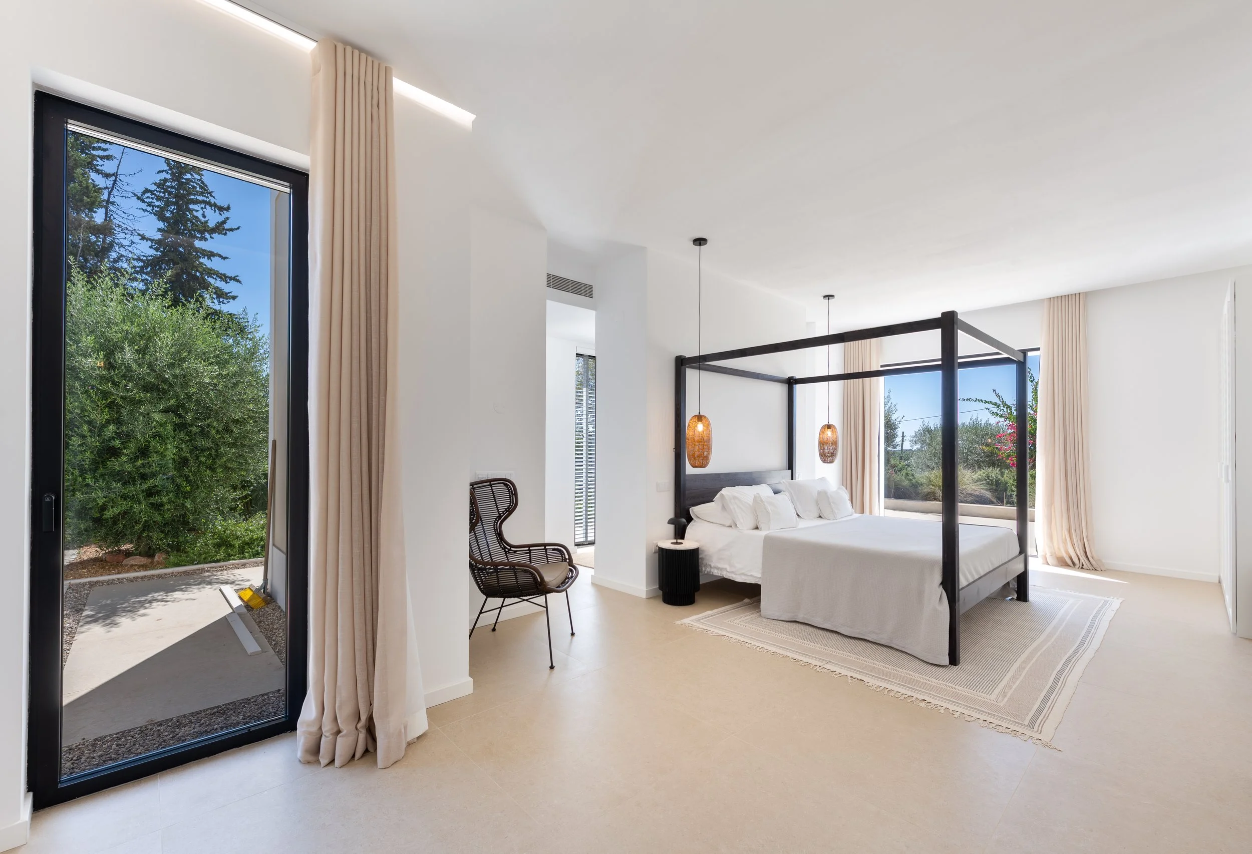 Quarto moderno com cama de casal, dois pendentes de luz de fibras e enfeite de cabeceira de madeira escura, piso claro, cortinas claras e grande janela com vista para natureza.