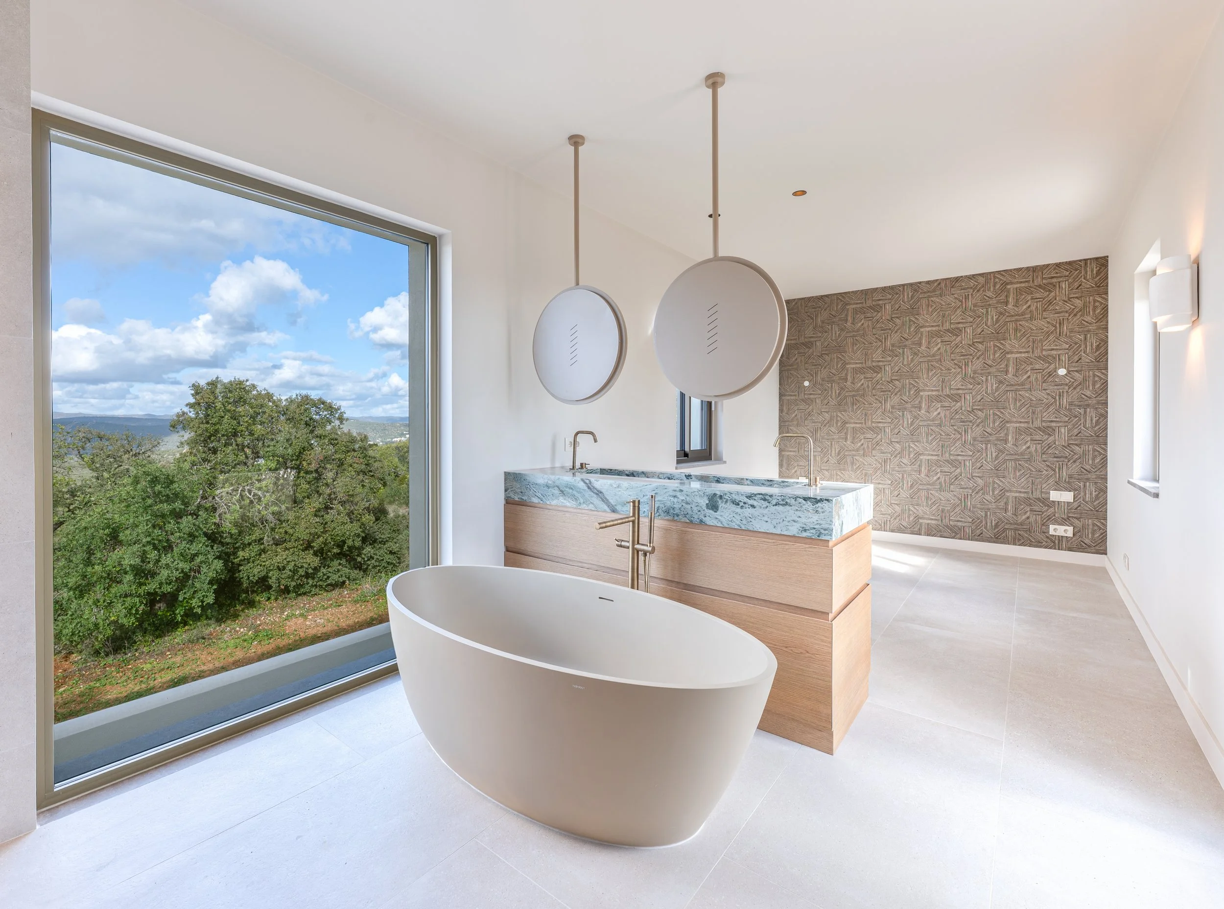 Banheira de spa com vista para a natureza, parede de azulejos decorativos na parede do fundo, janela grande com vista para o céu e árvores, iluminação suave na parede ao lado.