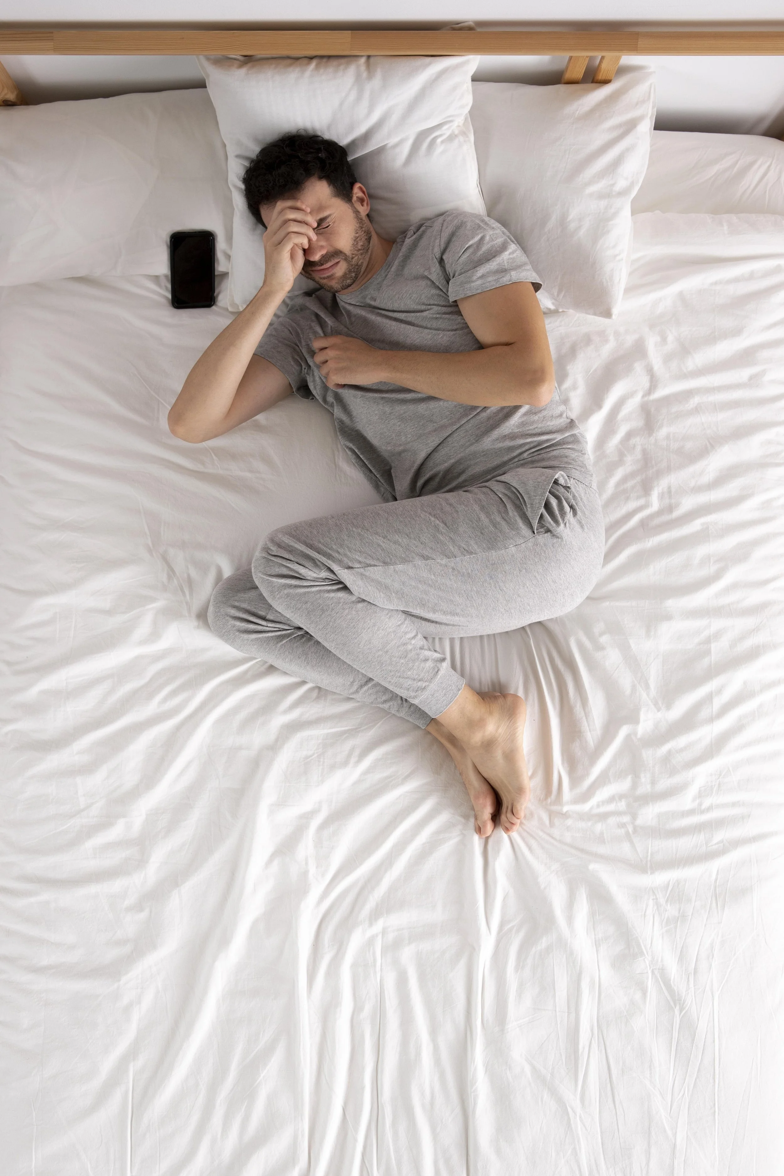 Un homme allongé dans un lit, semblant avoir mal à la tête ou être malade, avec un téléphone posé à côté de lui.