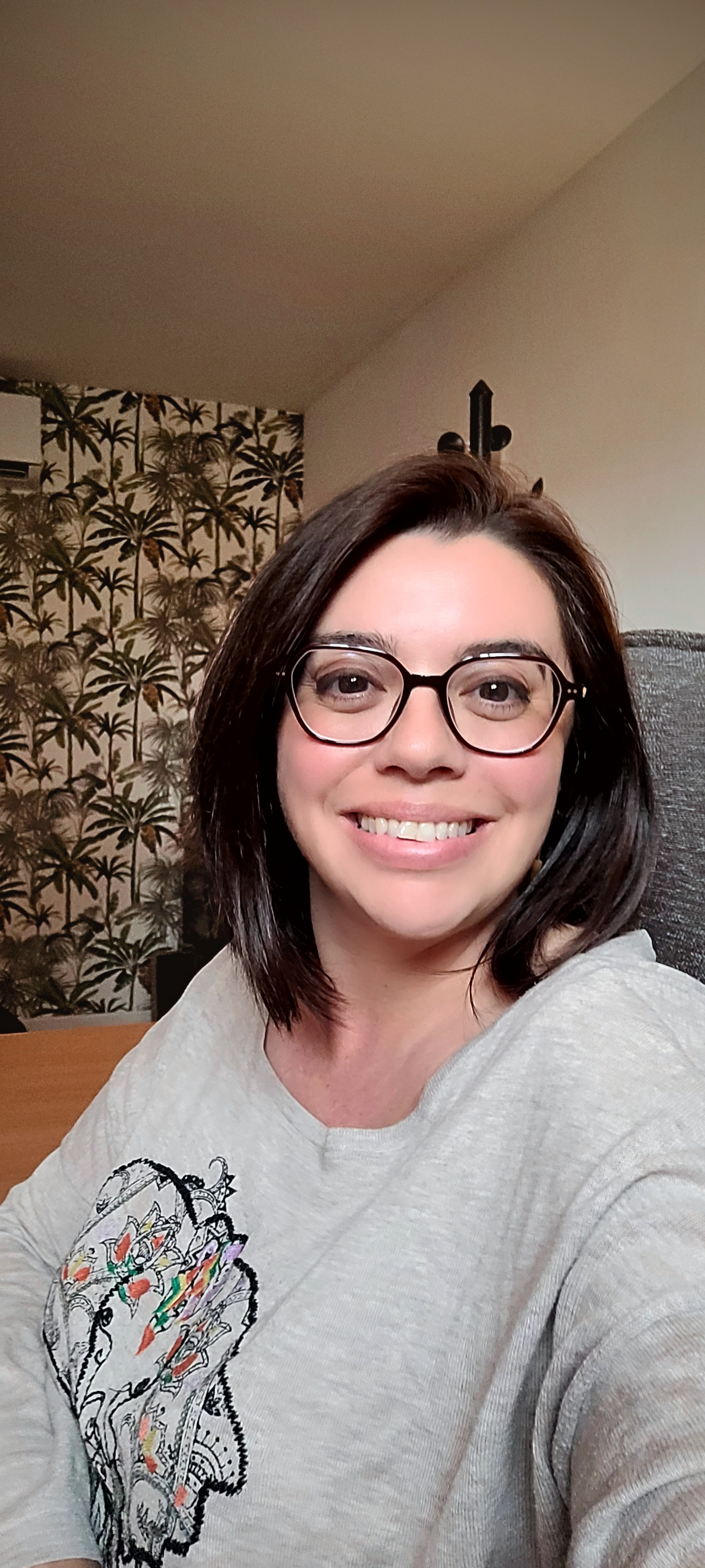 Une femme souriante avec des lunettes, portant un sweatshirt gris, assise dans une pièce avec un mur décoré de papier peint à motifs de palmiers.