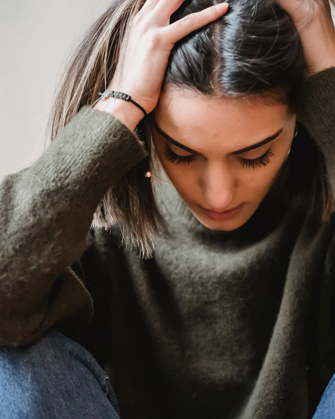 Une jeune femme avec les yeux fermés, tenant sa tête avec ses mains, portant un pull gris foncé et une boucle d'oreille pendante, semblant être en réflexion ou en détresse.