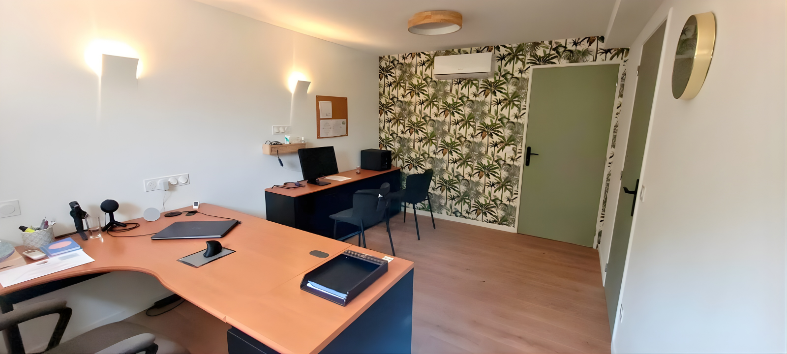 Bureau avec bureau principal, deux écrans, et une tablette, et un deuxième bureau avec deux chaises, un mur avec papier peint tropical, sol en bois, porte verte, et miroir mural.