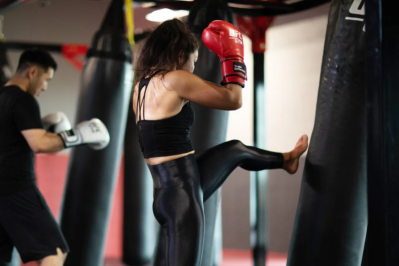 A woman wearing black leggings and a black sports top boxing with red gloves during a training session at a gym, with a man in the background also training with boxing gloves.