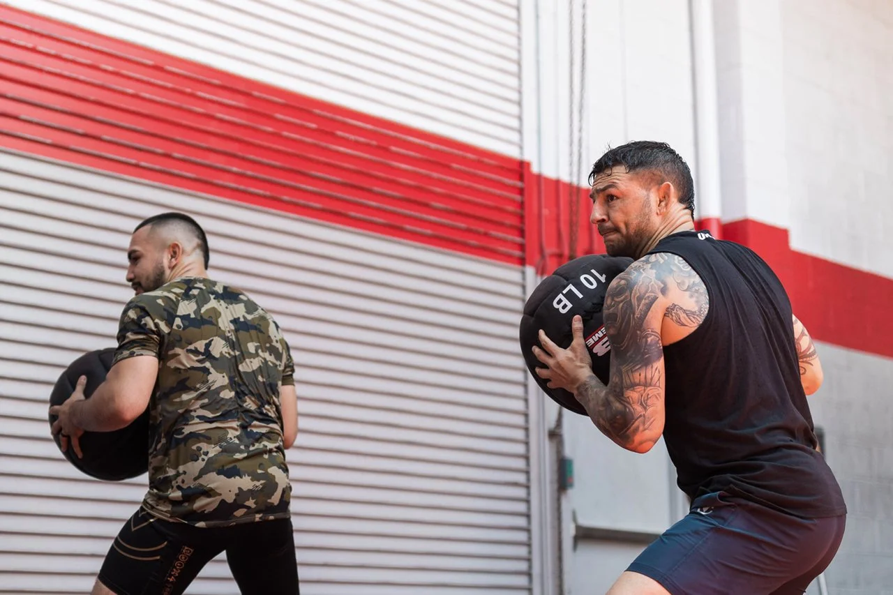 Two men lifting medicine ball in an industrial gym with red and white striped wall.