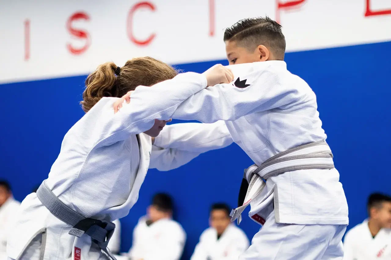 Two young girls competing in a judo match, wearing white gis with black belts, on a blue mat at a judo competition.