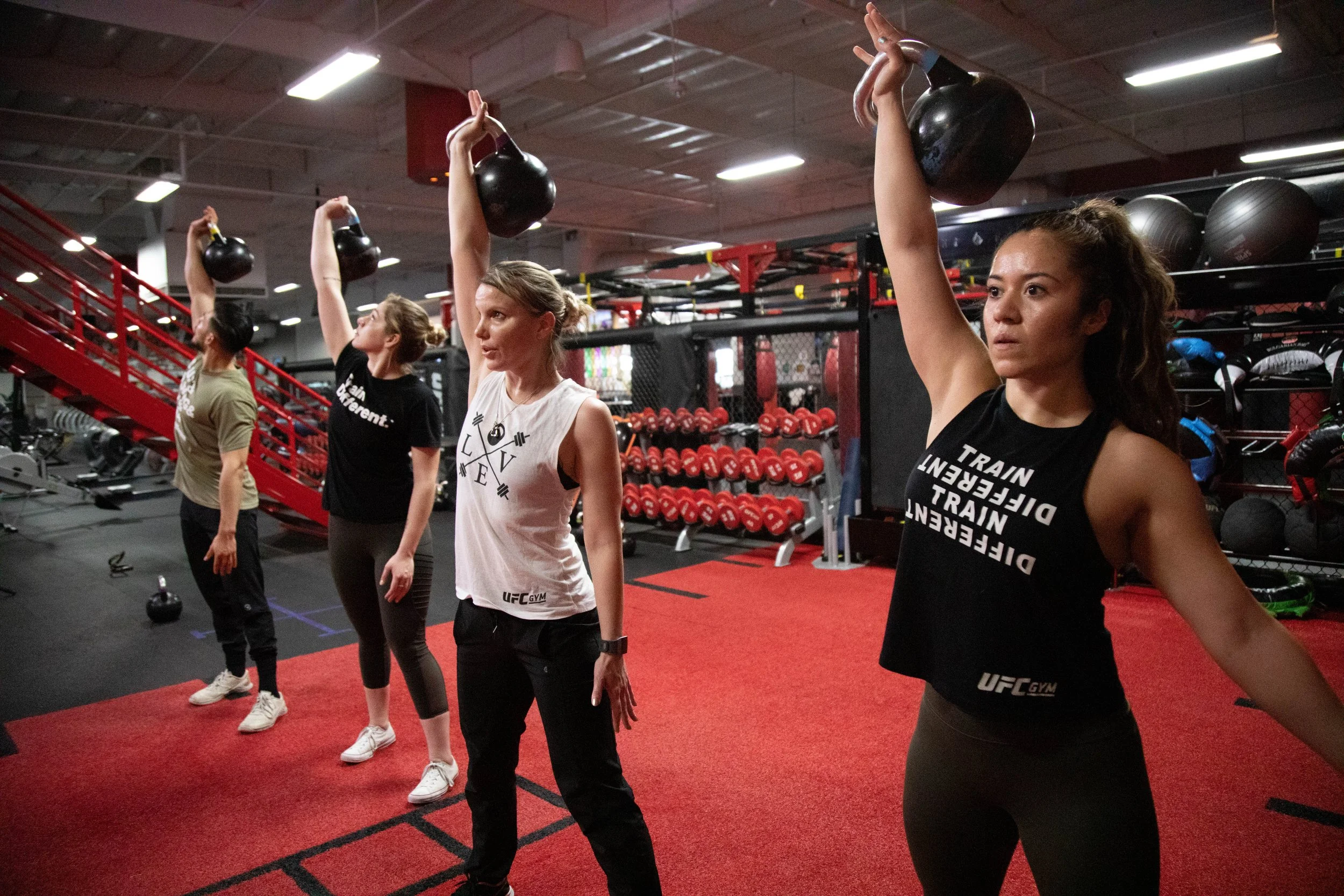 A group of women exercising with kettlebells in a gym, lifting kettlebells overhead during a workout session.