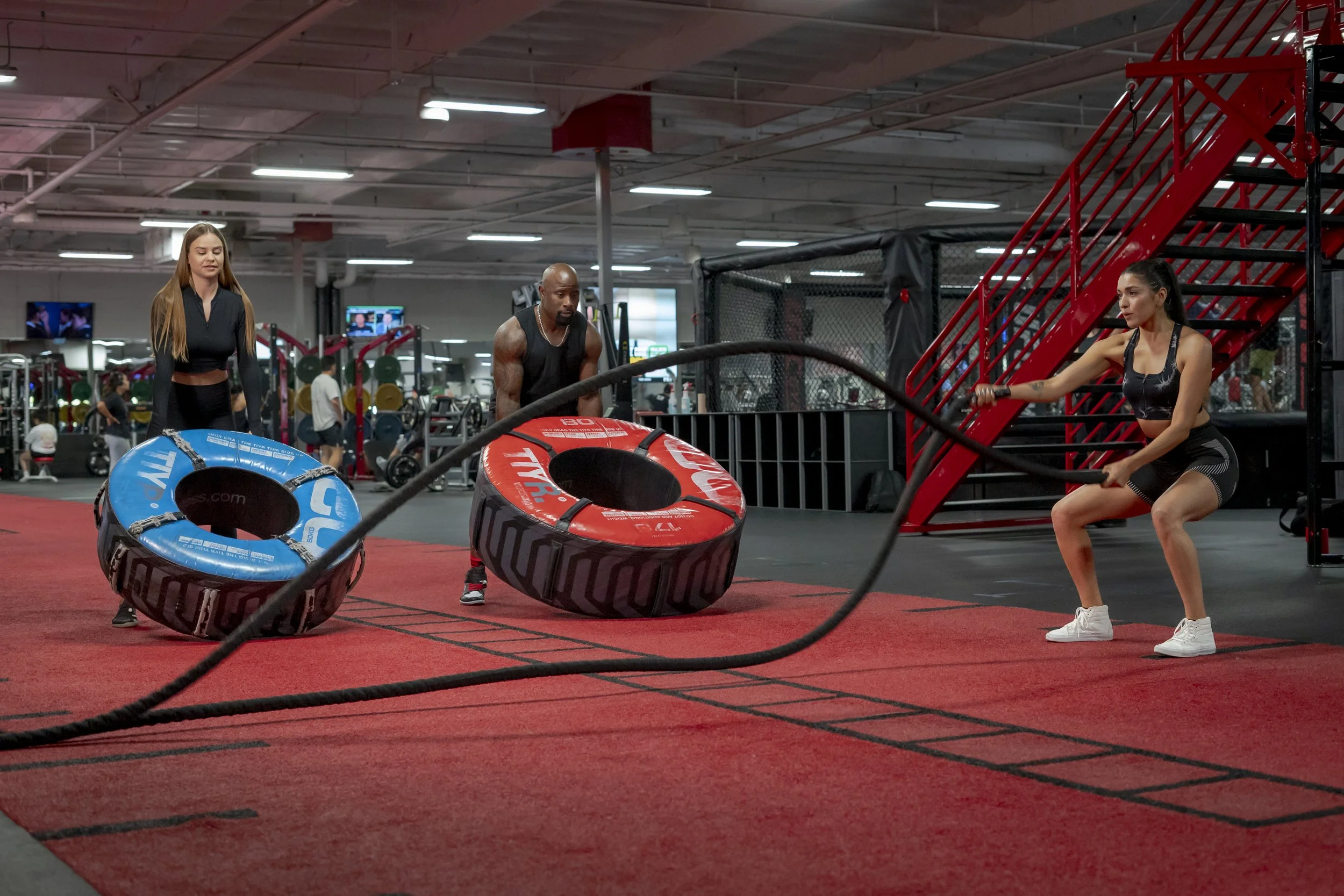 People working out in a gym with sleds, tires, and a staircase for training.