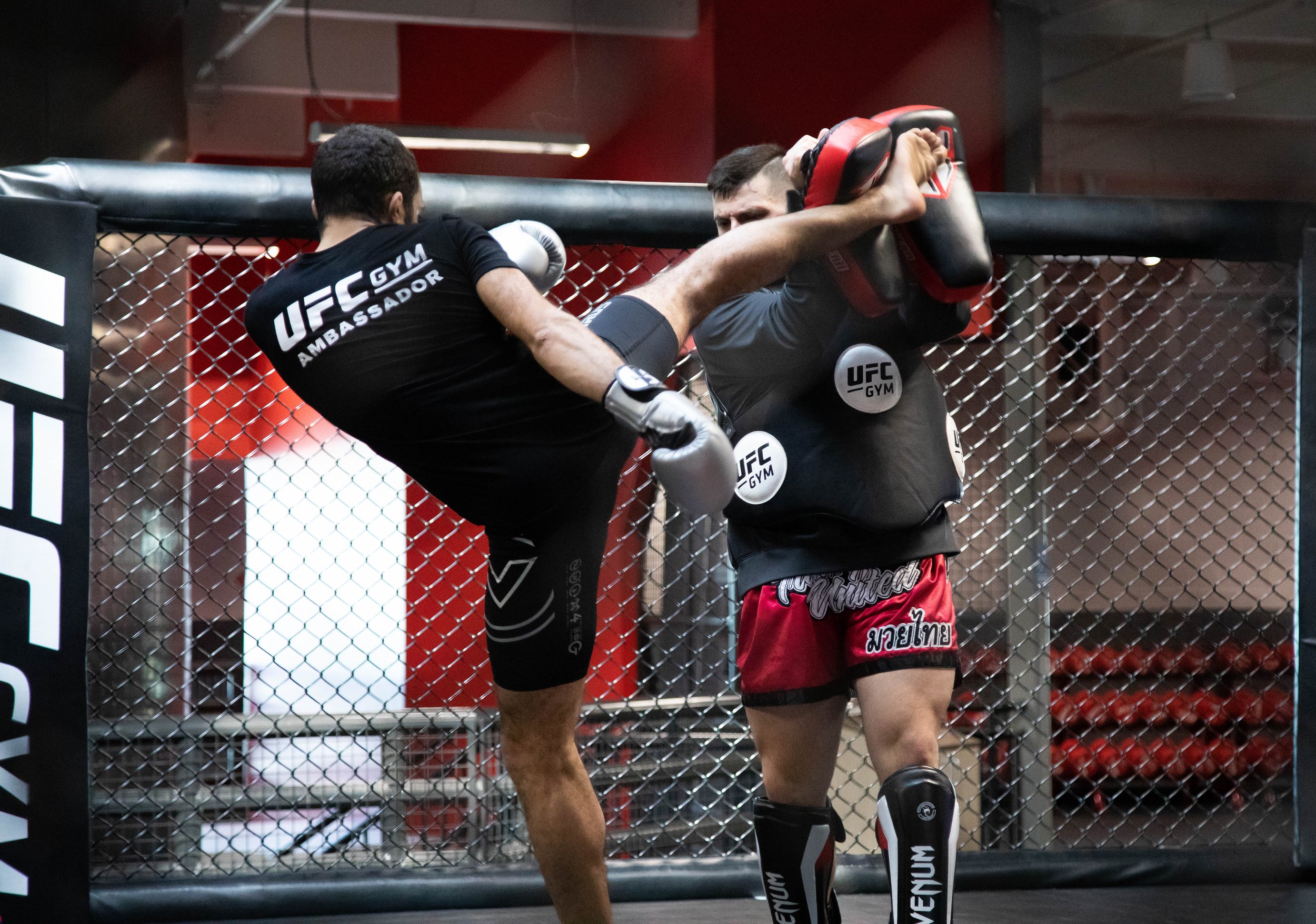 Two fighters engaged in a mixed martial arts match inside an octagon-shaped cage, with one fighter delivering a kick to the other.