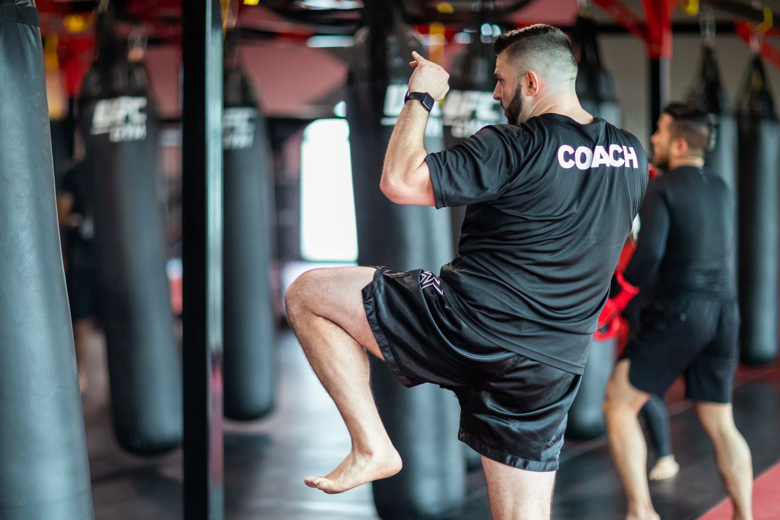A man with short hair and a beard stretching his leg on a balance training platform in a gym, with punching bags and other people exercising in the background.