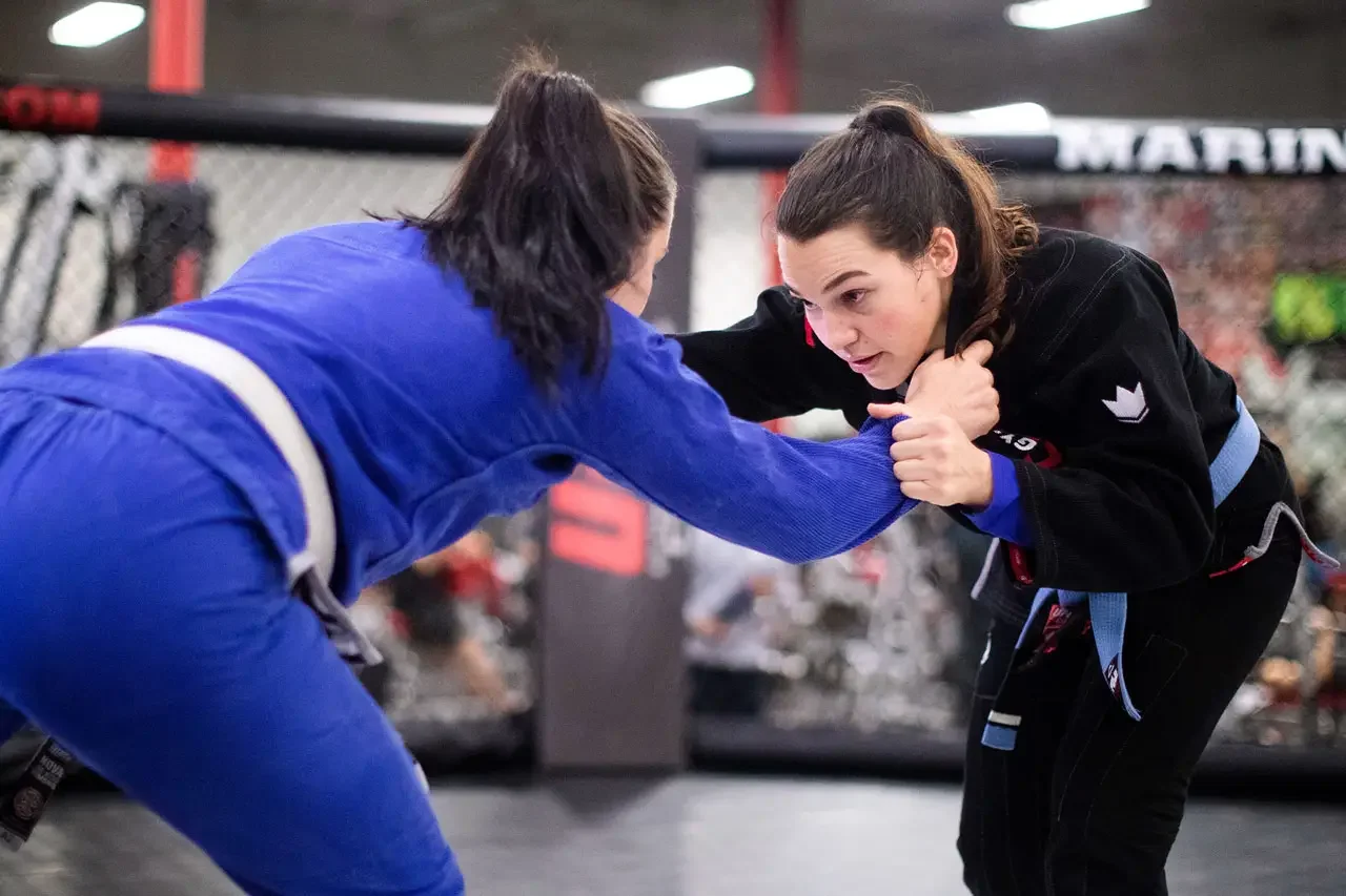 Two women practicing Brazilian Jiu-Jitsu in a fighting cage, one in a blue gi and the other in a black gi, engaging in a grappling hold.