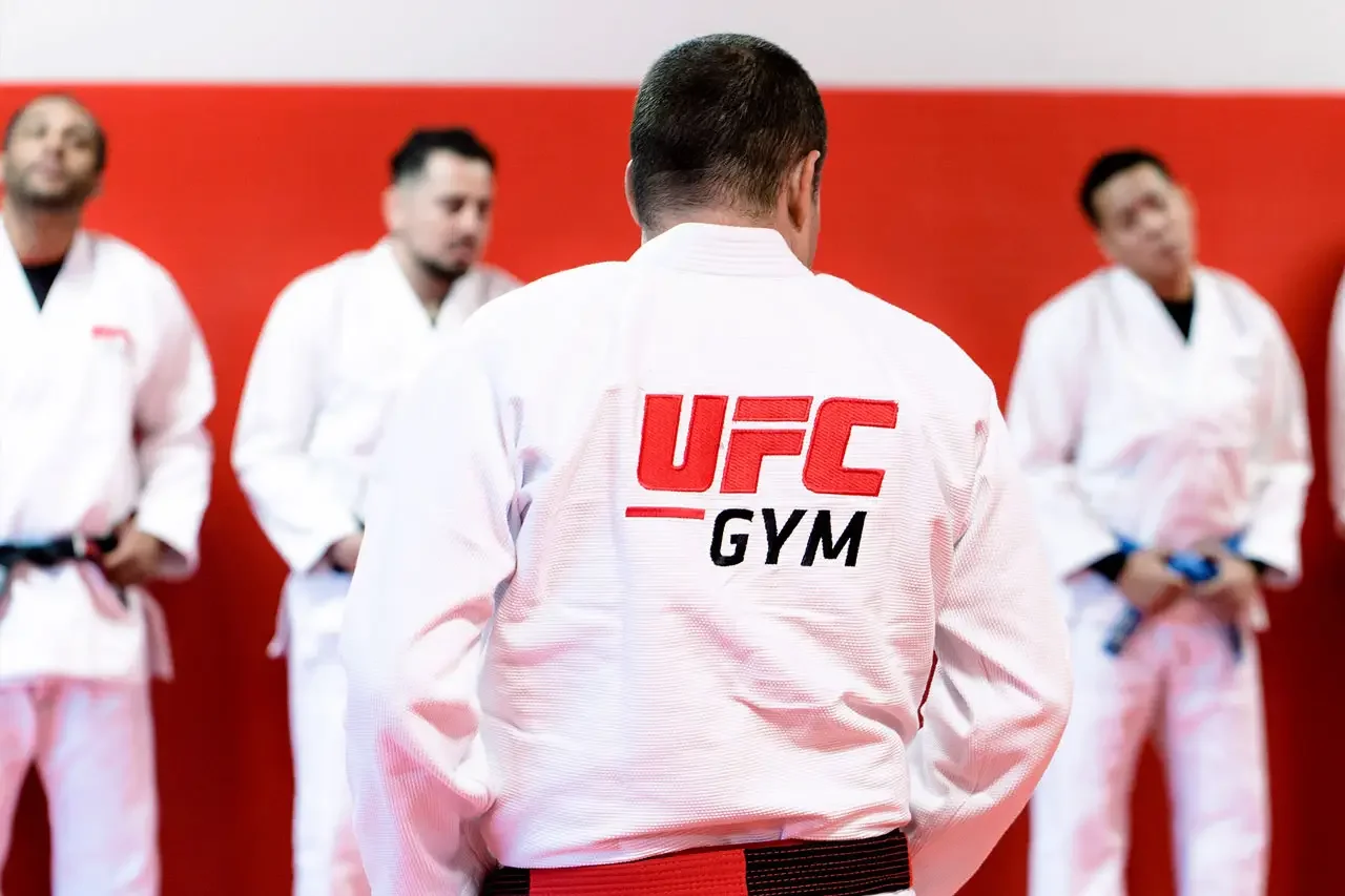 Martial arts students wearing gi uniforms, one with a UFC GYM logo on the back, standing in a dojo with a red background.