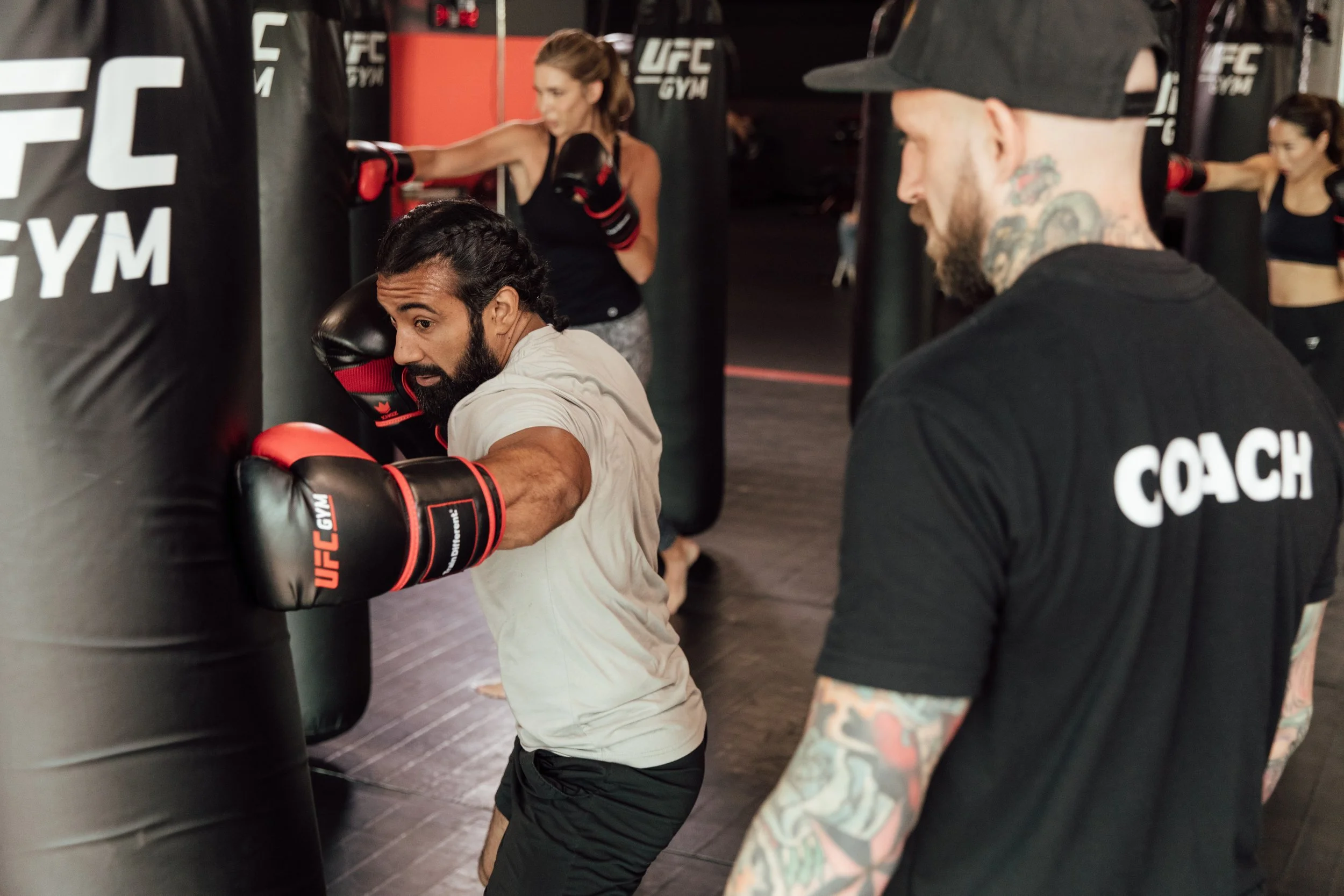A person with a beard and cornrows is practicing boxing at a gym, punching a black punching bag with red gloves. A coach, wearing a black T-shirt with white lettering and a cap, observes closely. Two women in workout attire can be seen in the backgro