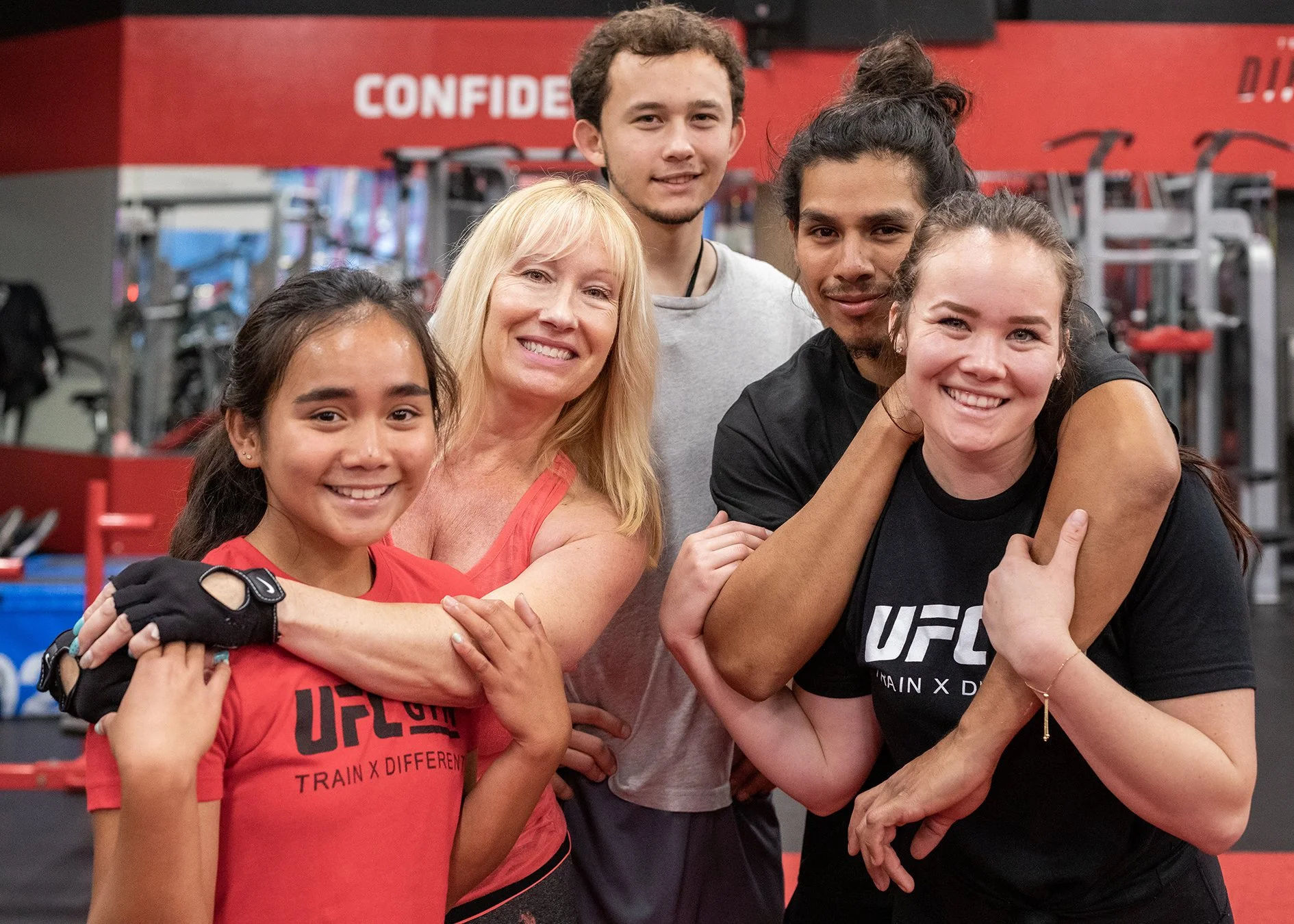 A group of five people, including a young girl, a woman, and three young adults, smiling and hugging in a gym with red walls and workout equipment. Some are wearing UFC branded workout shirts.