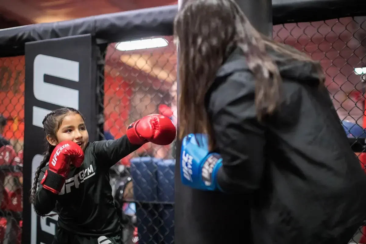 Young girl in boxing gloves practicing her punches on her coach inside a UFC gym training cage.
