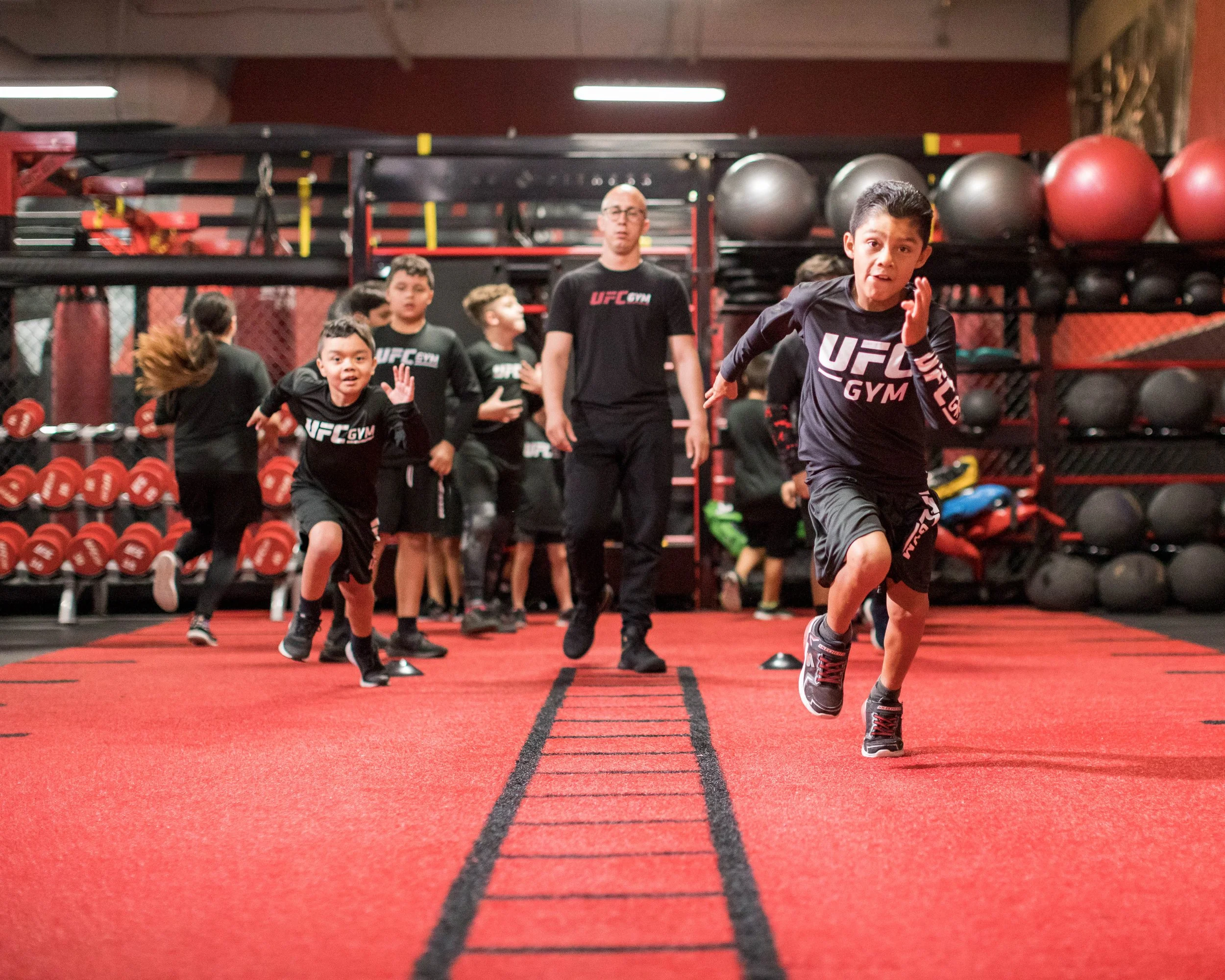 Children and an adult running in a gym, with red flooring and equipment in the background.