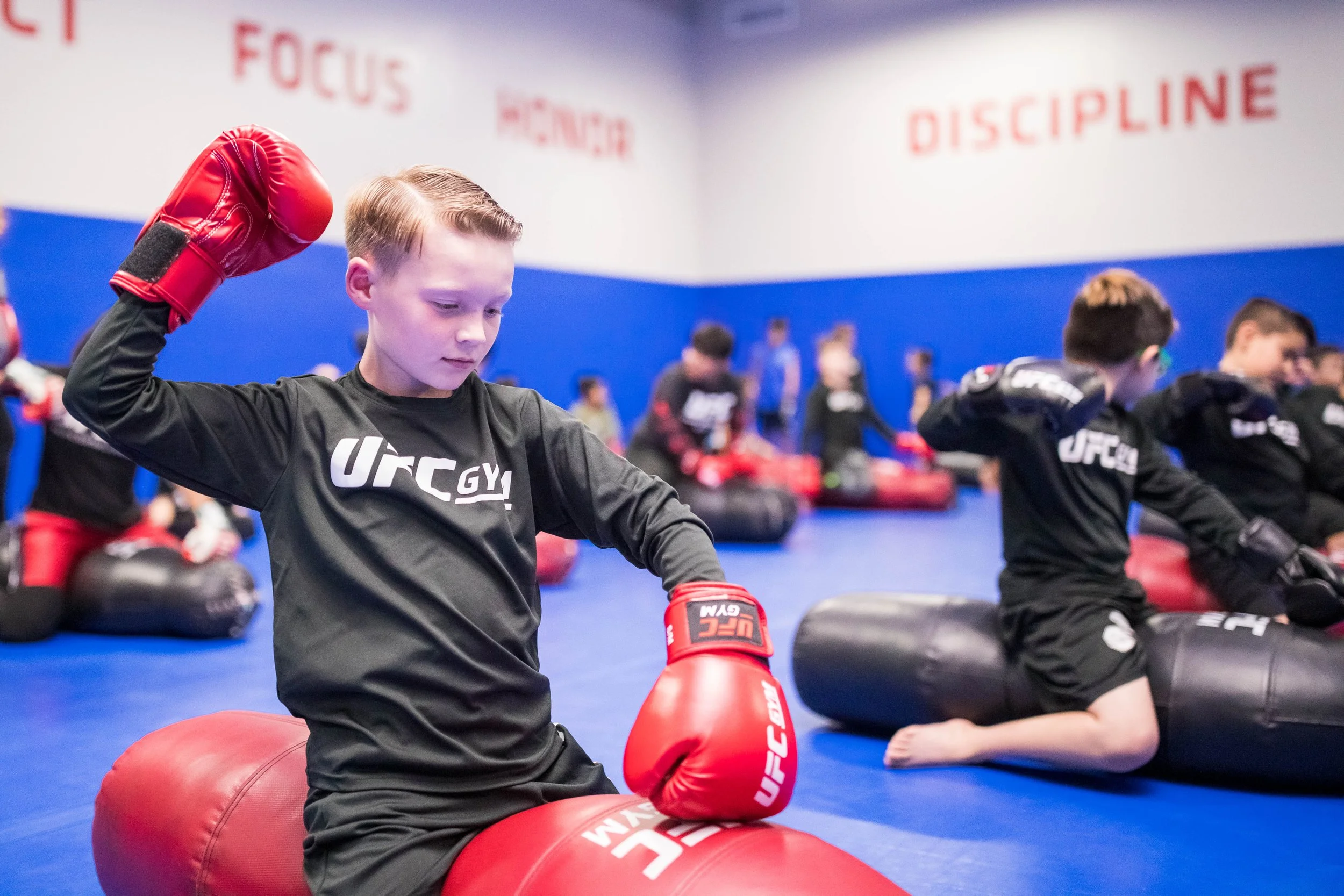 Young boys in black martial arts uniforms sitting on blue mats in a training room, practicing boxing with red boxing gloves and punching bags in front of them. The room has white and blue walls with motivational words written on them in red, such as 'FOCUS' and 'DISCIPLINE'.