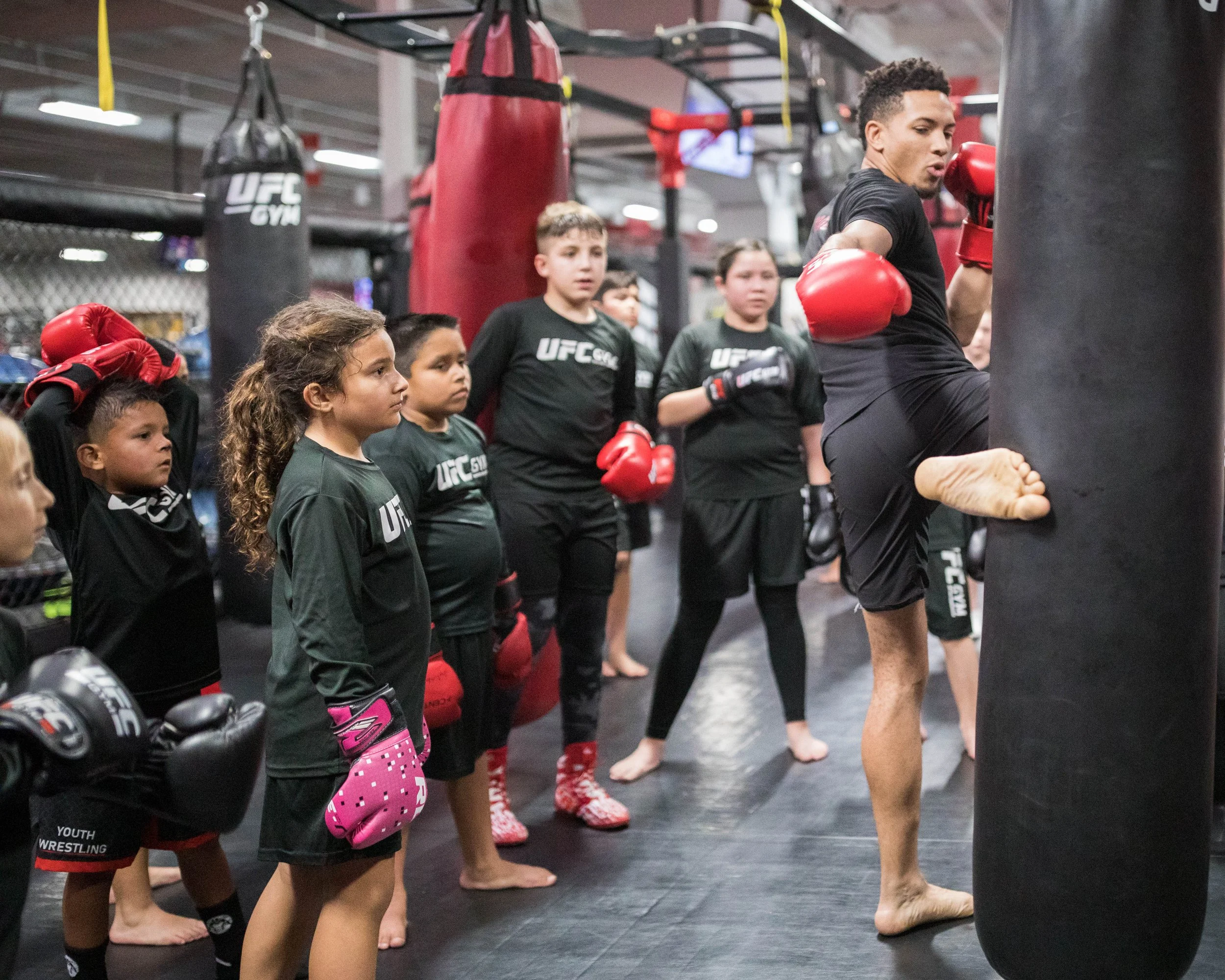 Young children and an adult practicing boxing in a gym, wearing black UFC Youth Wrestling shirts, red boxing gloves, and standing in a line near punching bags.