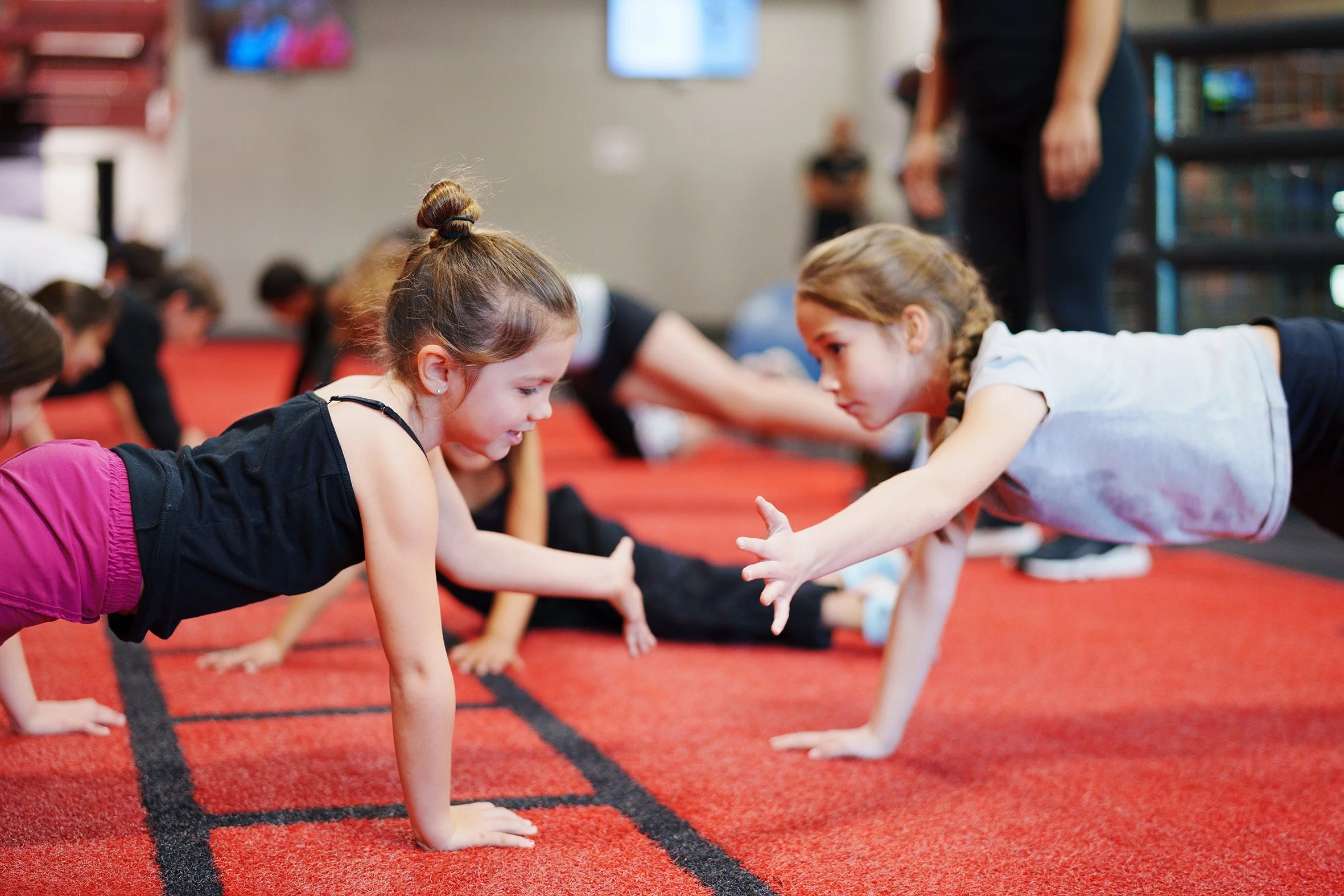 Two young girls engaging in a hand-on-hand activity on a red carpeted floor in a gym or classroom setting, with other children and an instructor in the background.