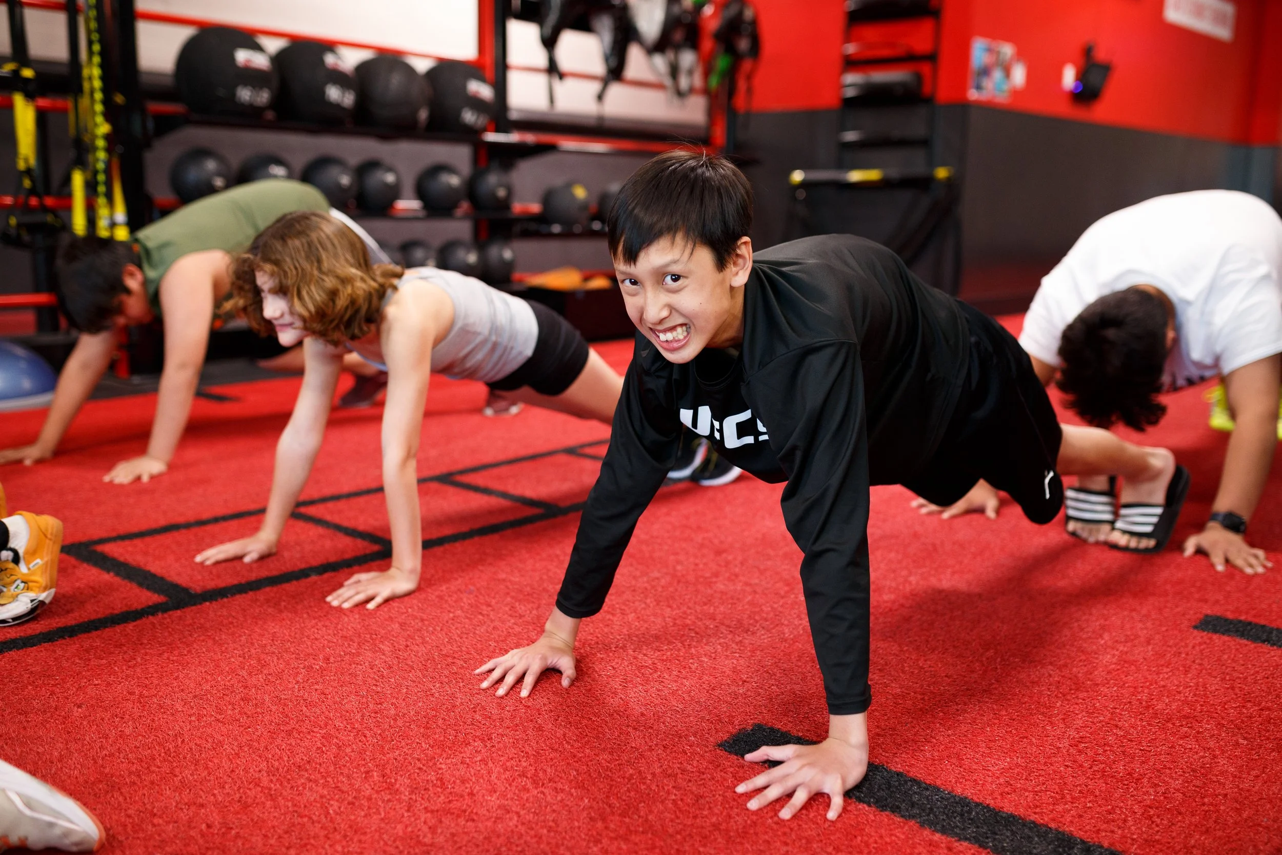 Group of children doing push-ups on red gym floor inside fitness gym with medicine balls and workout equipment in background.