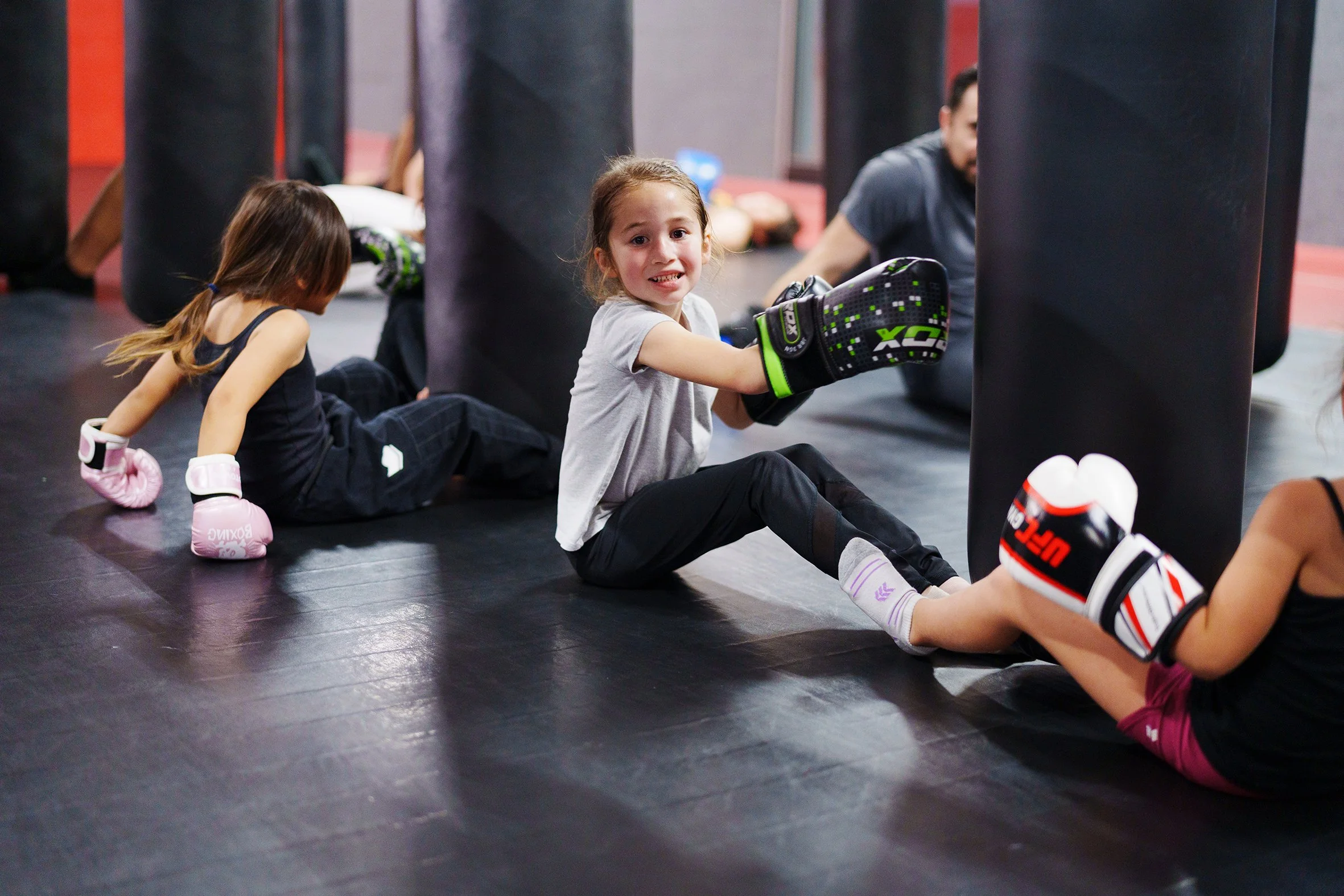 Children participating in a boxing class, sitting on the floor with punching bags and boxing gloves in a gym.