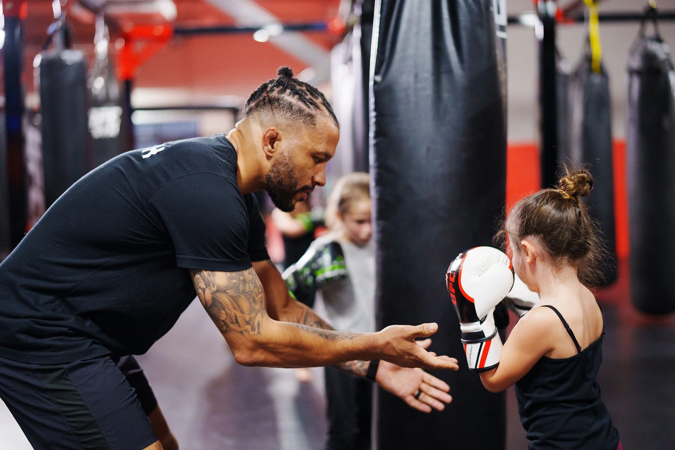 An adult man with tattooed arms and braided hair coaching a young girl in boxing inside a gym, with punching bags in the background.