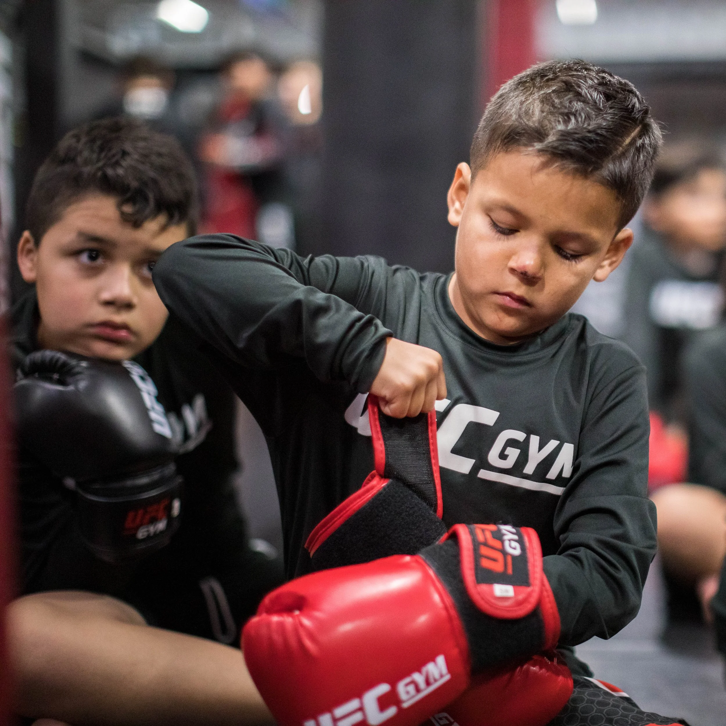 Two young boys wearing boxing gloves, one in black and red gloves and the other in red gloves, practicing boxing in a gym.