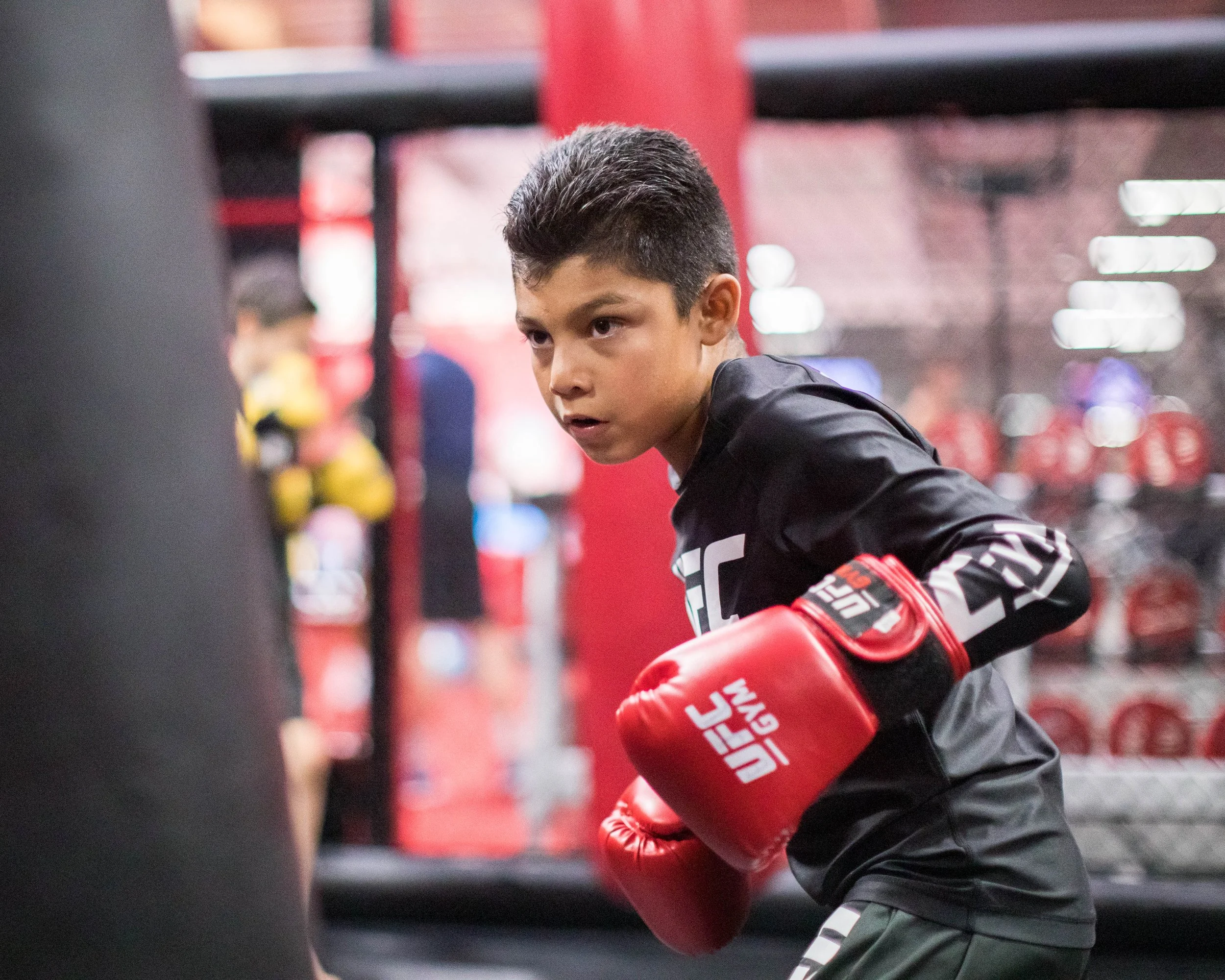 A young man with short dark hair wearing red boxing gloves and a black athletic shirt, practicing fitness drills in a gym, focused and determined.
