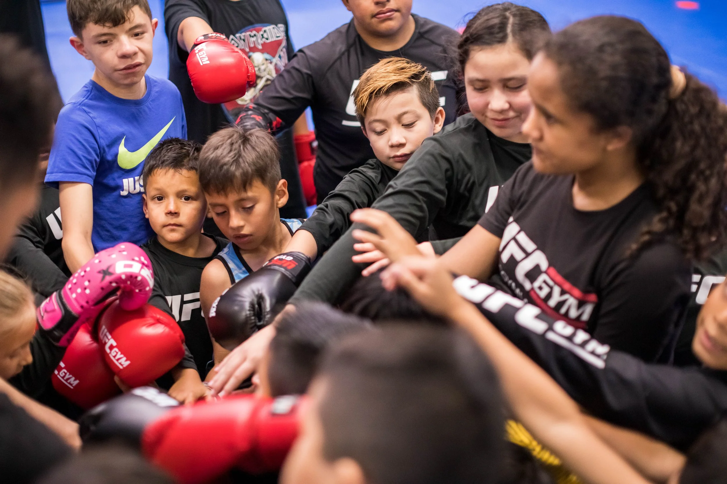 A group of young children and a woman gather closely together in a boxing gym, with some children wearing boxing gloves. The children appear to be listening attentively or preparing for a boxing session.