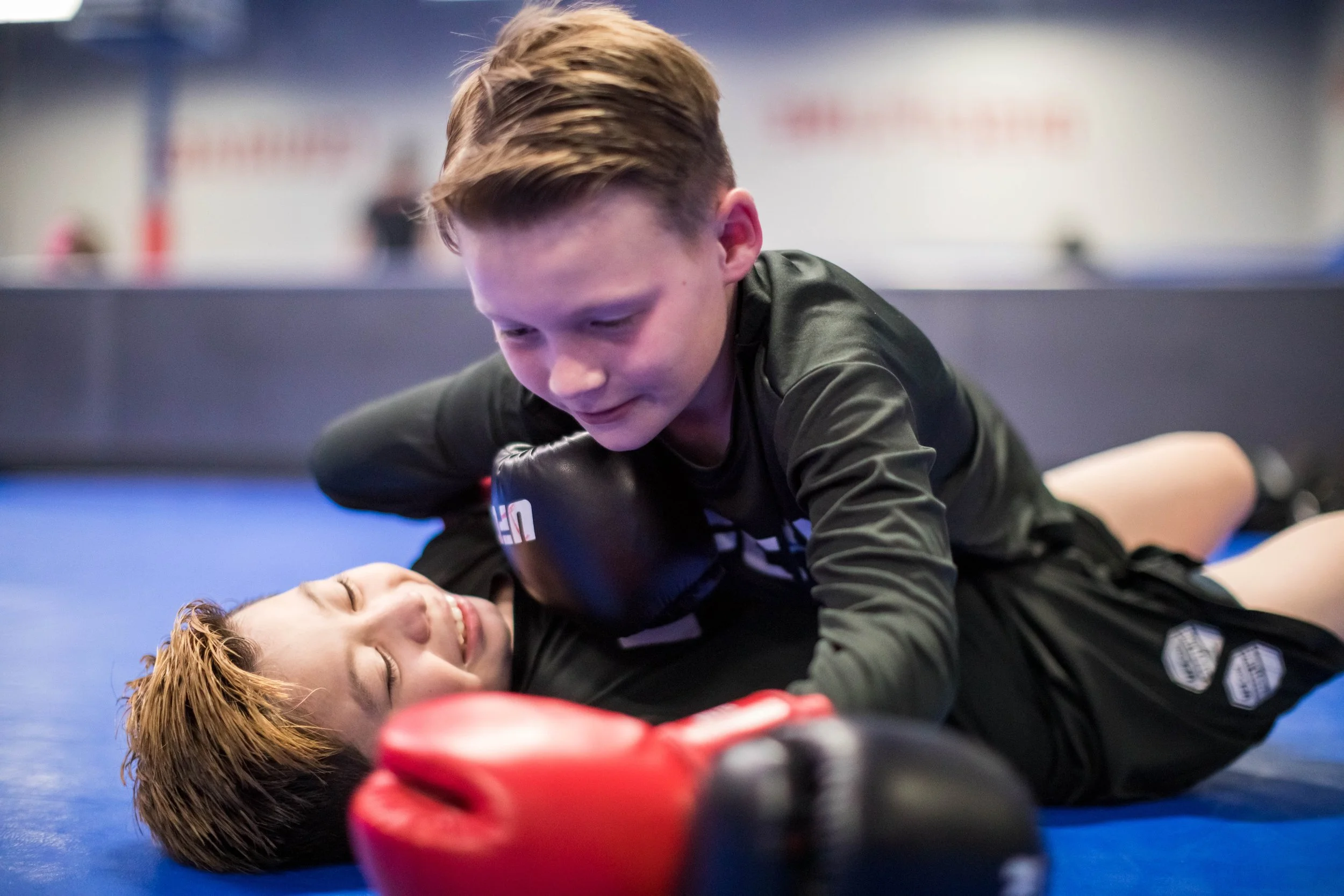 Two martial artists engaged in a grappling match on blue mats, with one on top and the other lying on her back, smiling and looking relaxed.